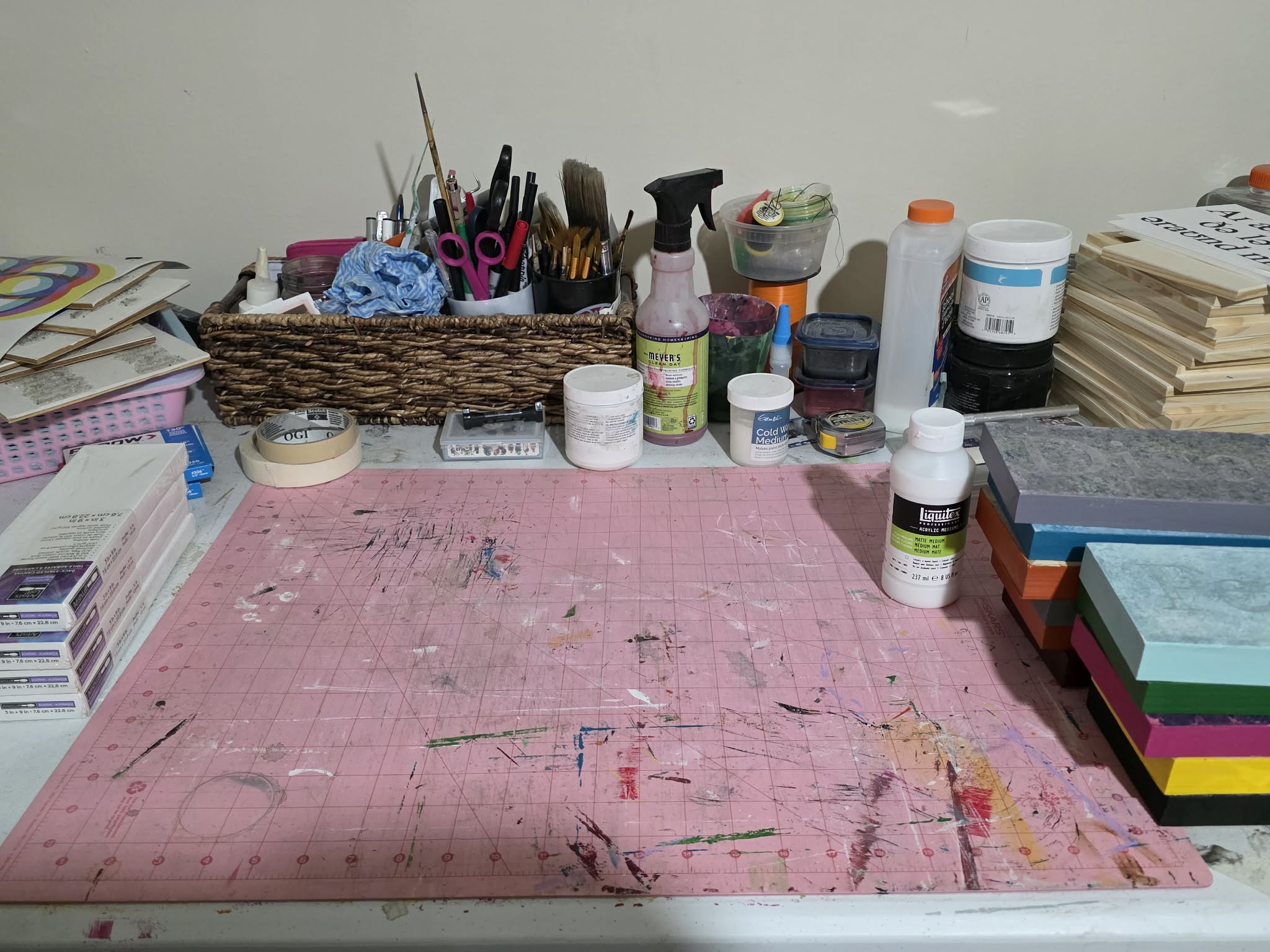 Work surface with art supplies, including paint bottles, brushes, and stacks of paper, on a pink table with stains.