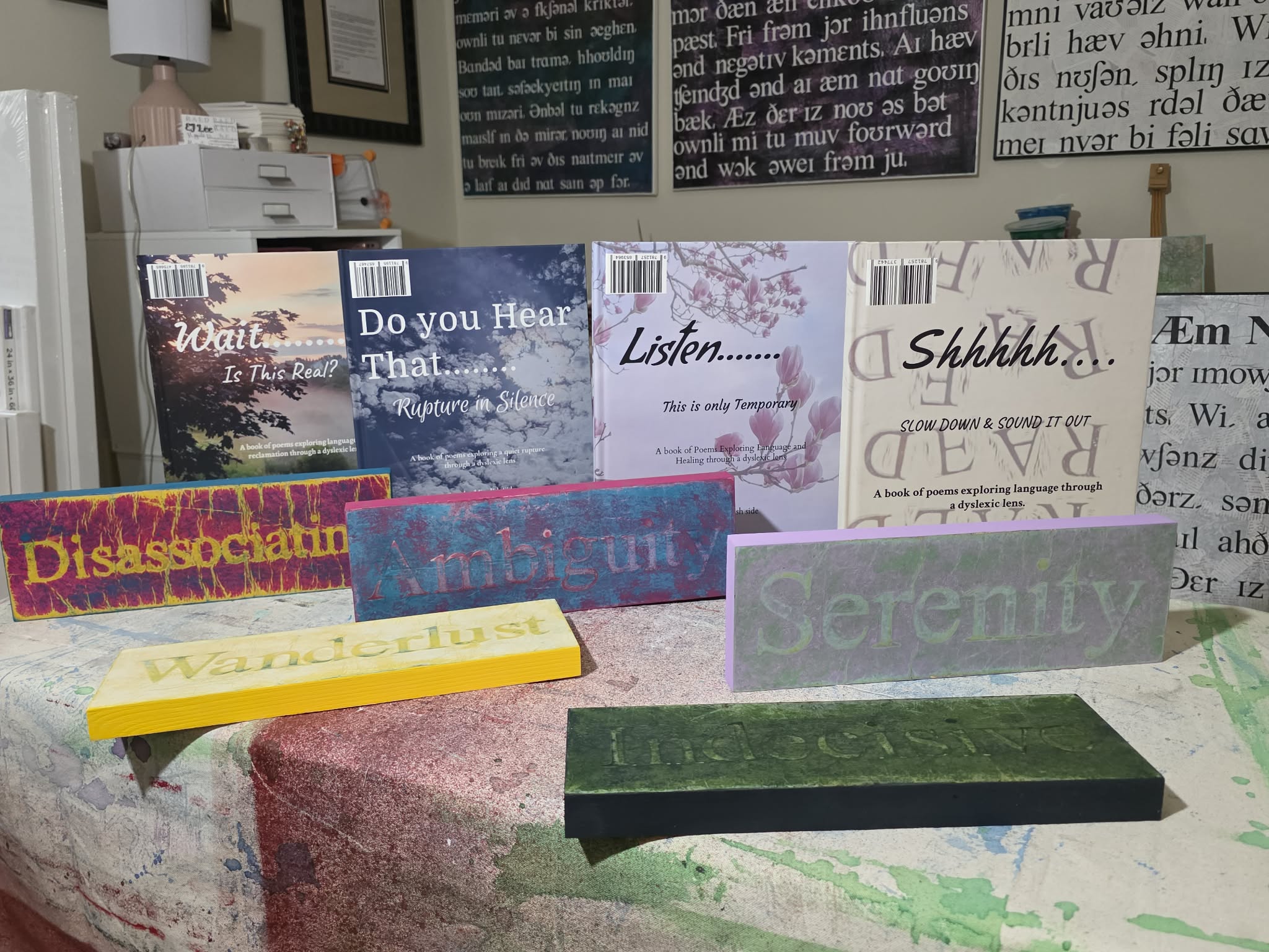 Display of various books and signs on a table, some with text like 'Disassociation,' 'Serenity,' and 'Listen,' with a pink table surface.