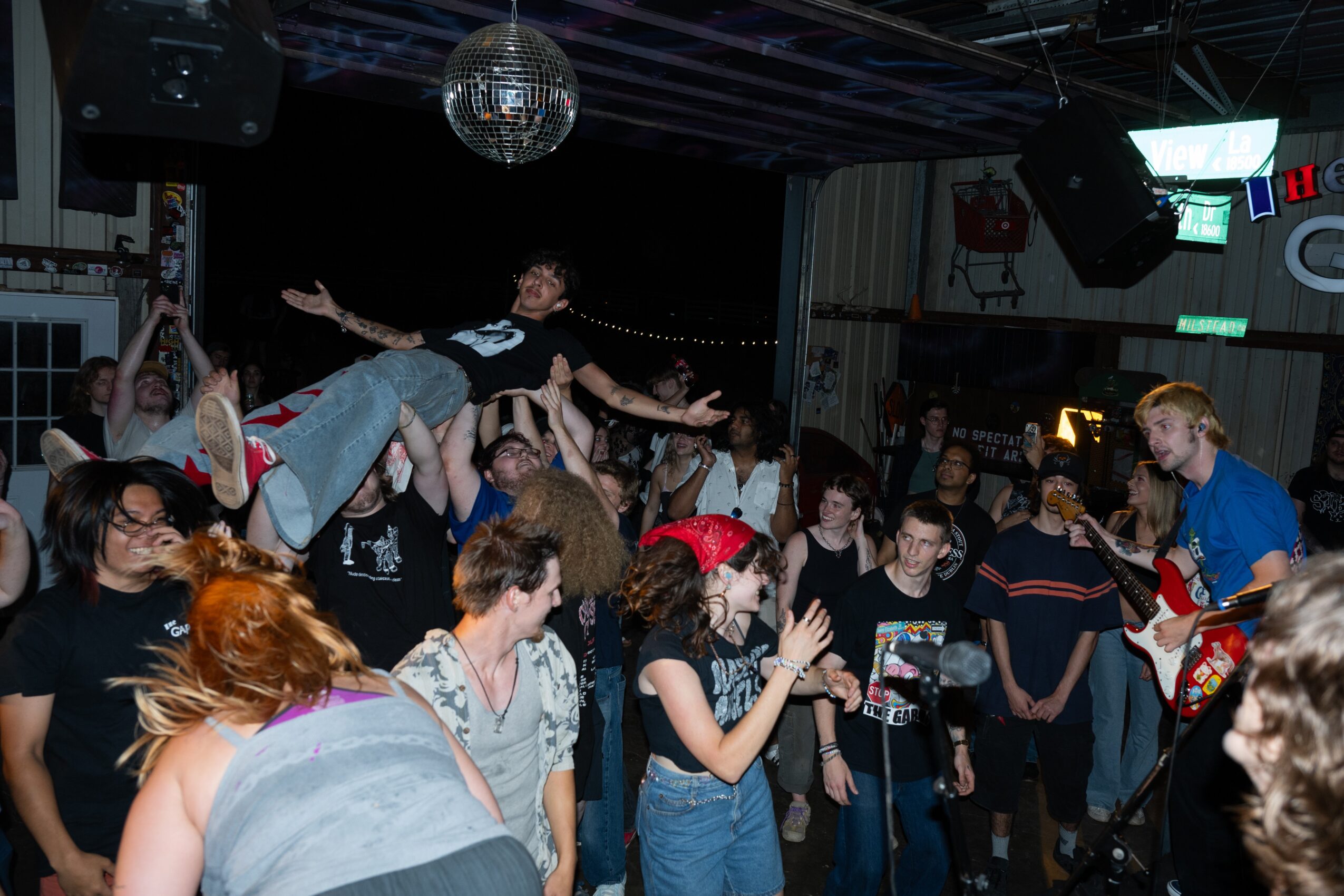 People gathered in a club with a person being lifted above the crowd, disco ball overhead, and a stage in the background.