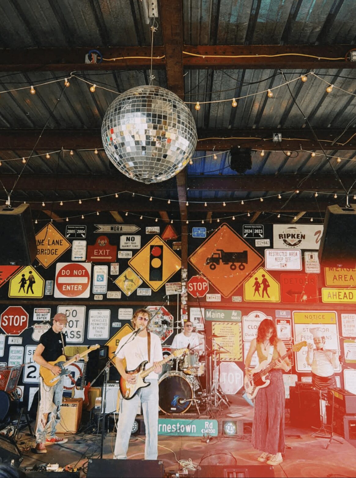 Band performing on stage with a disco ball hanging from ceiling, decorated with signs and string lights, in a rustic venue.
