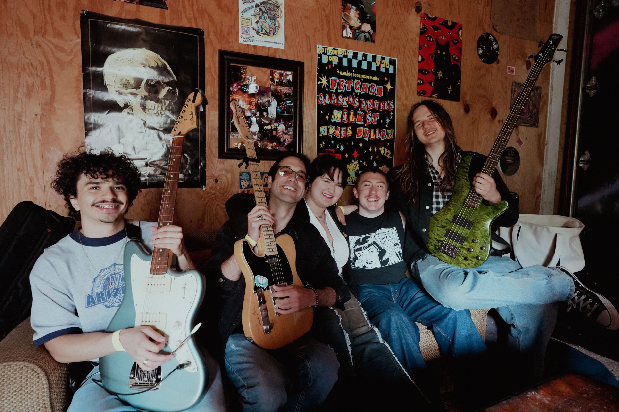 Five people sitting on a couch holding guitars in a room with wall art and a wooden wall background.