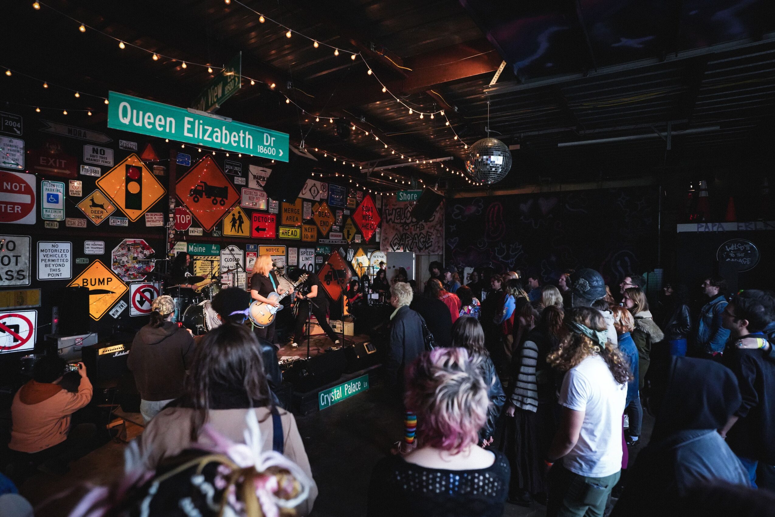 Crowd gathered in a dimly lit bar with colorful wall decorations and a stage, under a Queen Elizabeth street sign.