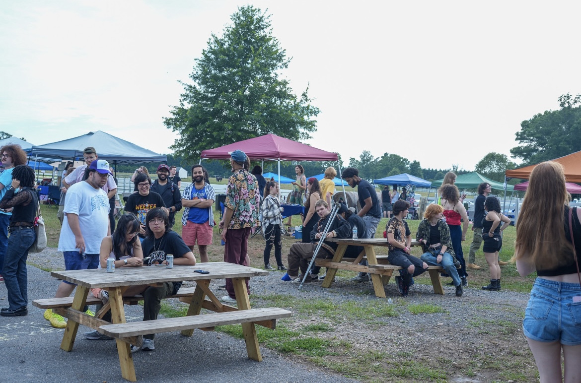 People gather outdoors around picnic tables and tents, some sitting and some standing, with trees and sky in background.
