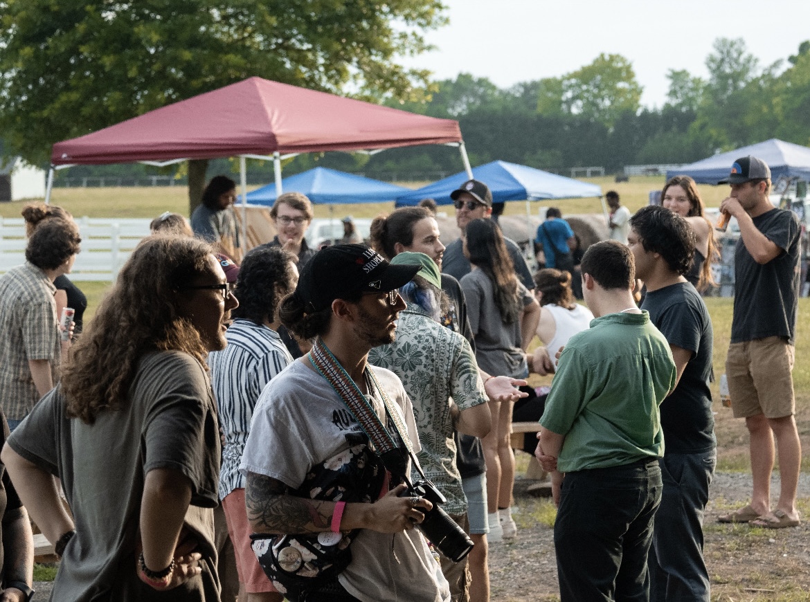Group of people gathered outdoors under tents, some talking and taking photos, with trees and a field in background.
