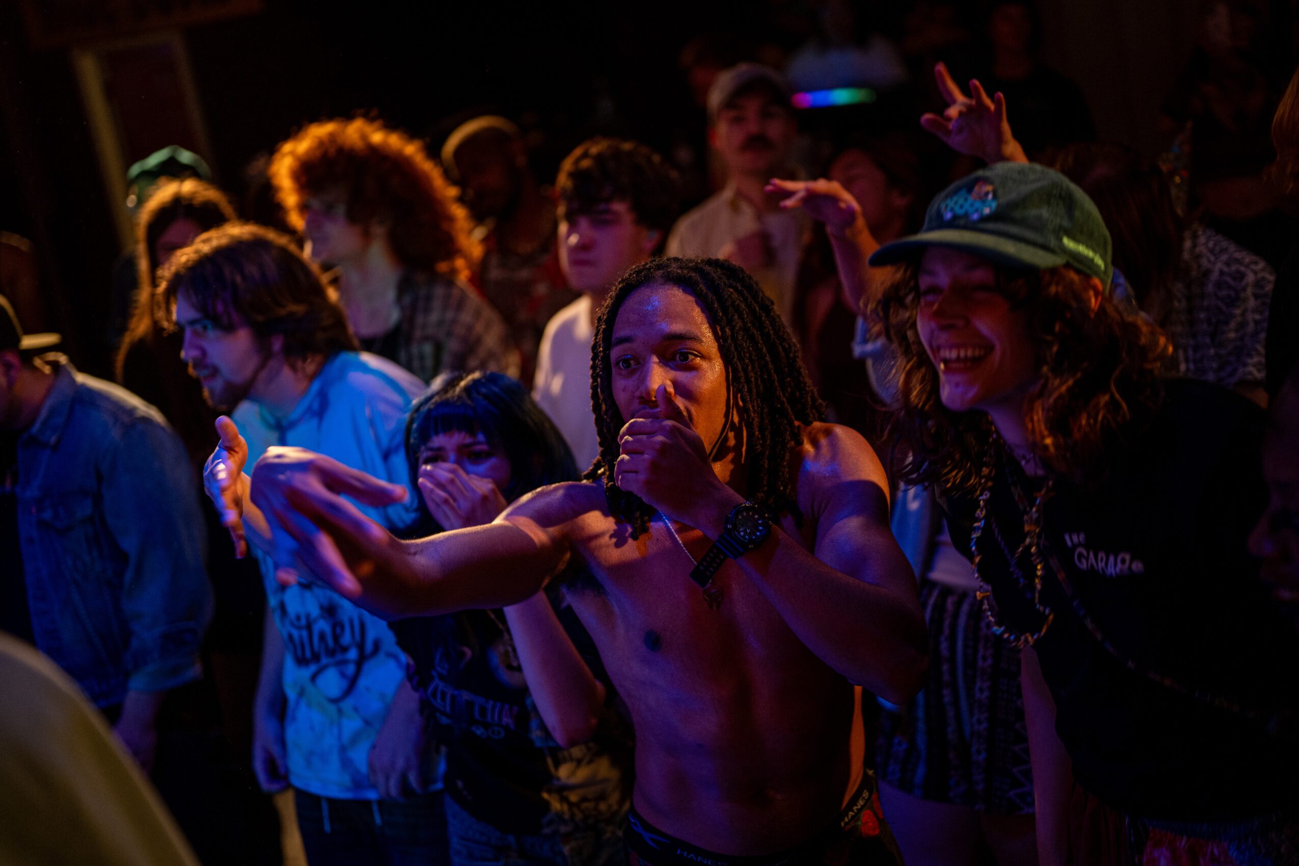 Group of people at a concert or event, some smiling and engaging with the performer, with colorful lighting in the background.