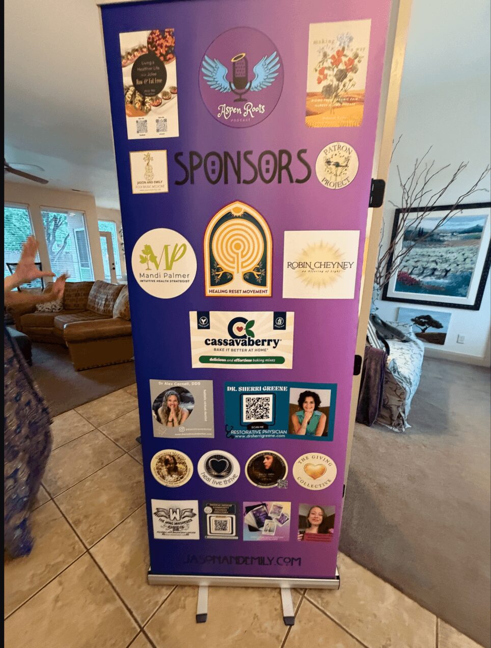 Purple sponsor display board with logos and images, placed indoors near a window and a couch, with a person taking a photo.