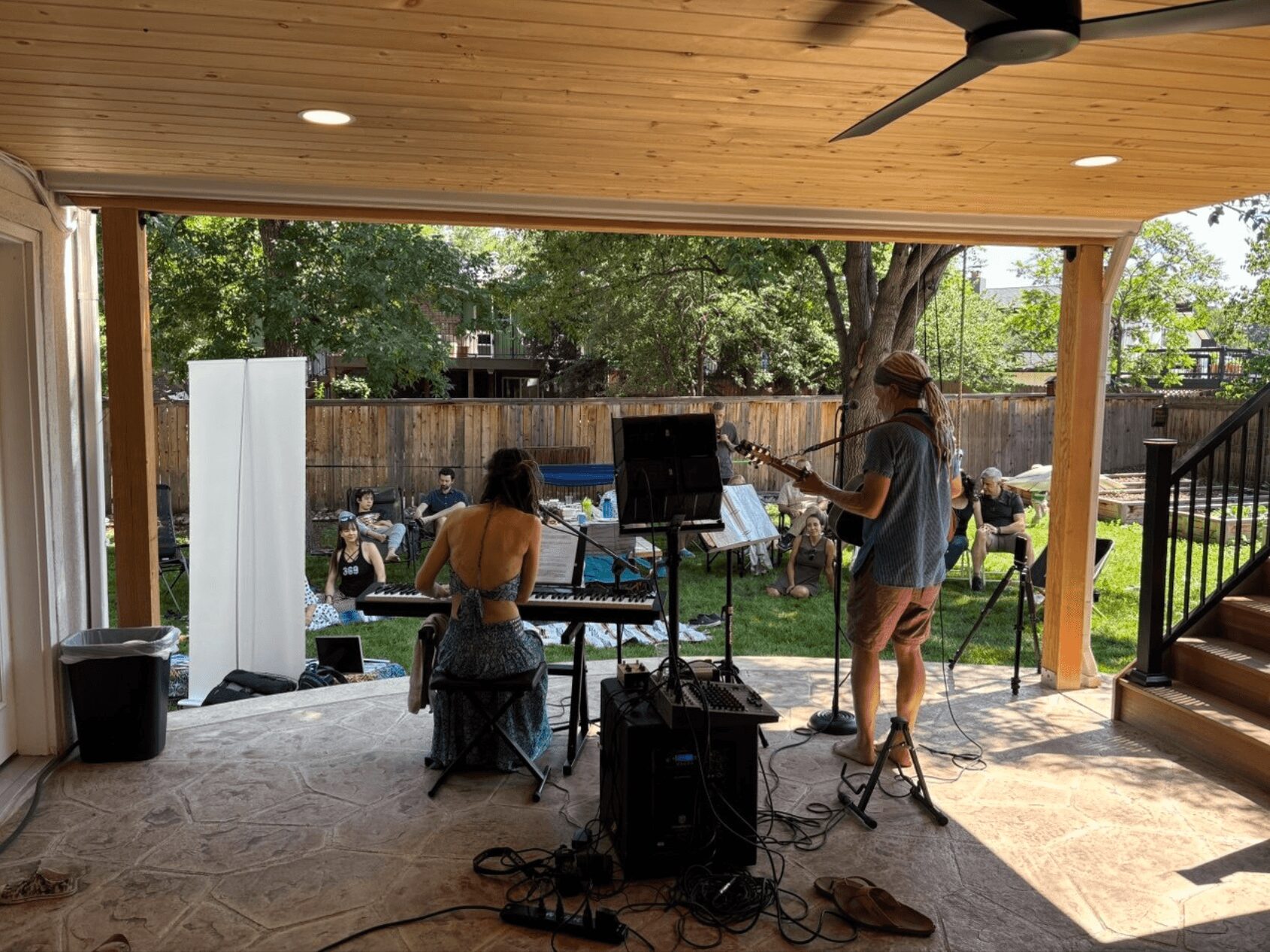 People performing music outdoors on a covered patio with a wooden ceiling, trees, and a backyard with seated guests.