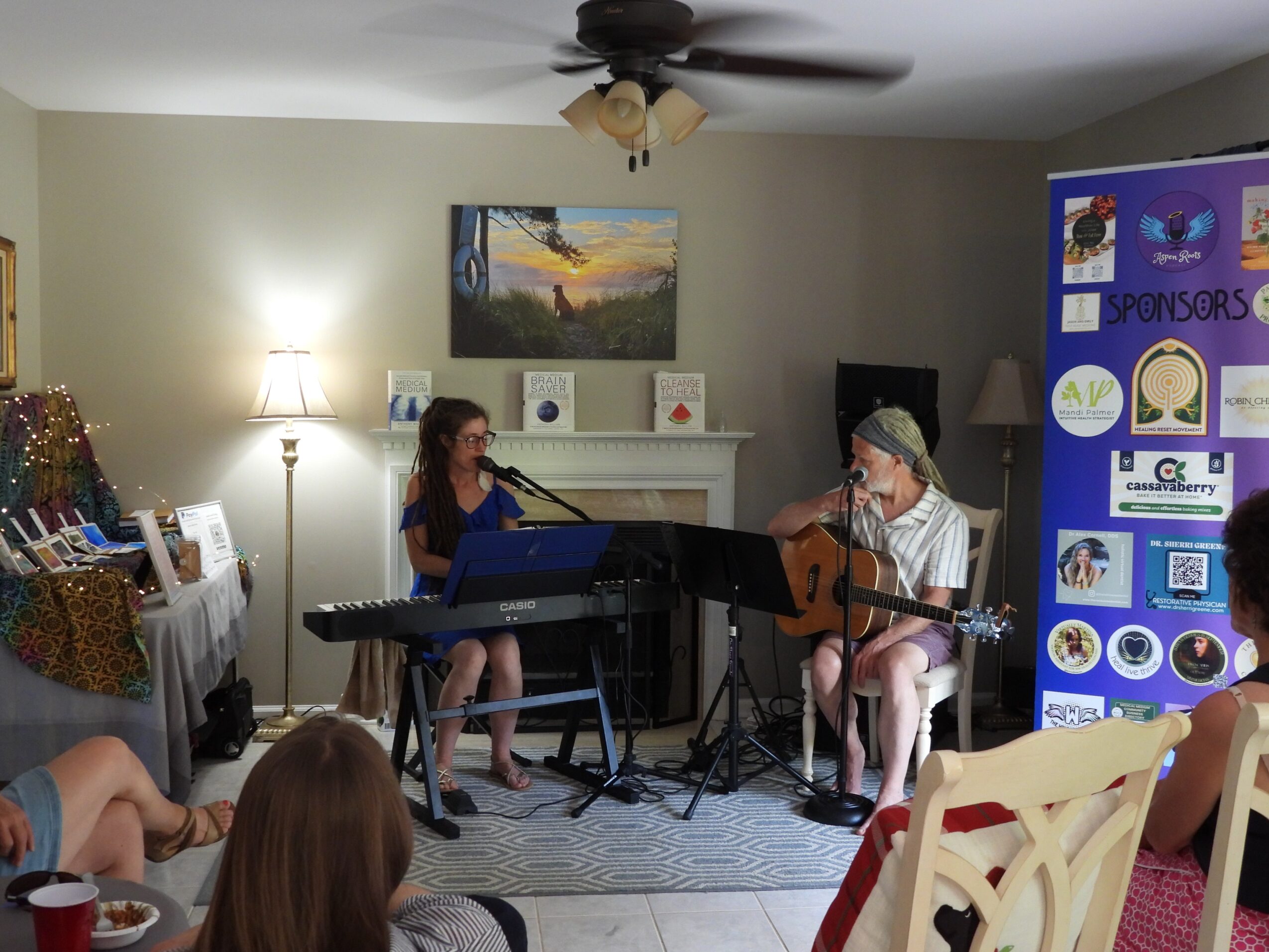 Two women perform music in a living room with a fireplace, piano, and audience, with a blue sponsor board on the right.