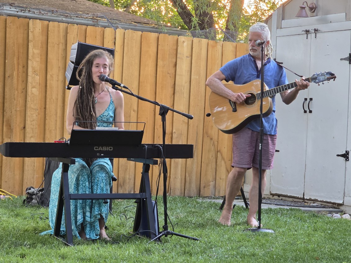 A young woman and an older man perform music outdoors, with a wooden fence and trees in the background.