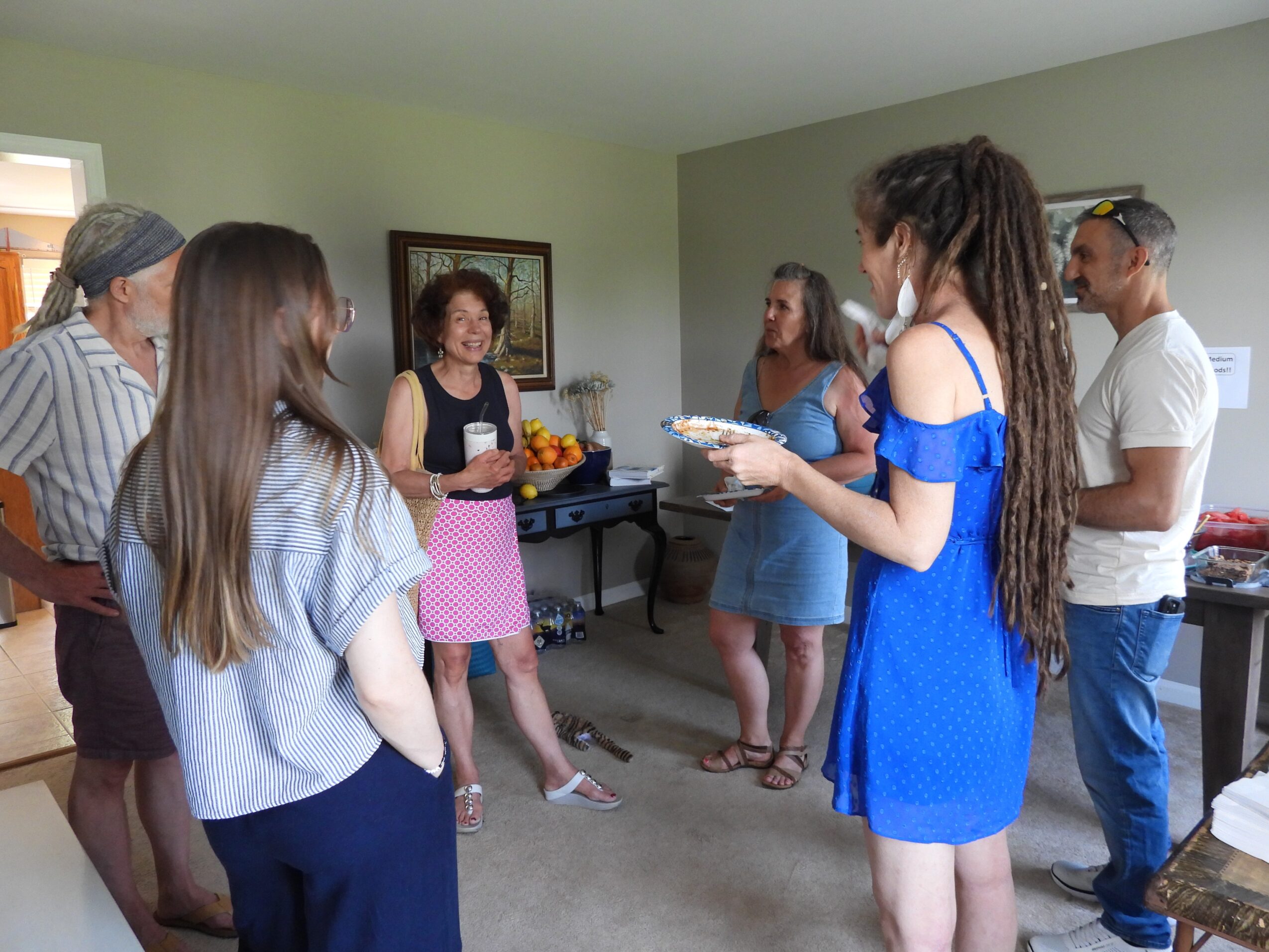 Group of seven people standing in a room, engaged in conversation, with a woman holding a plate of food, smiling.