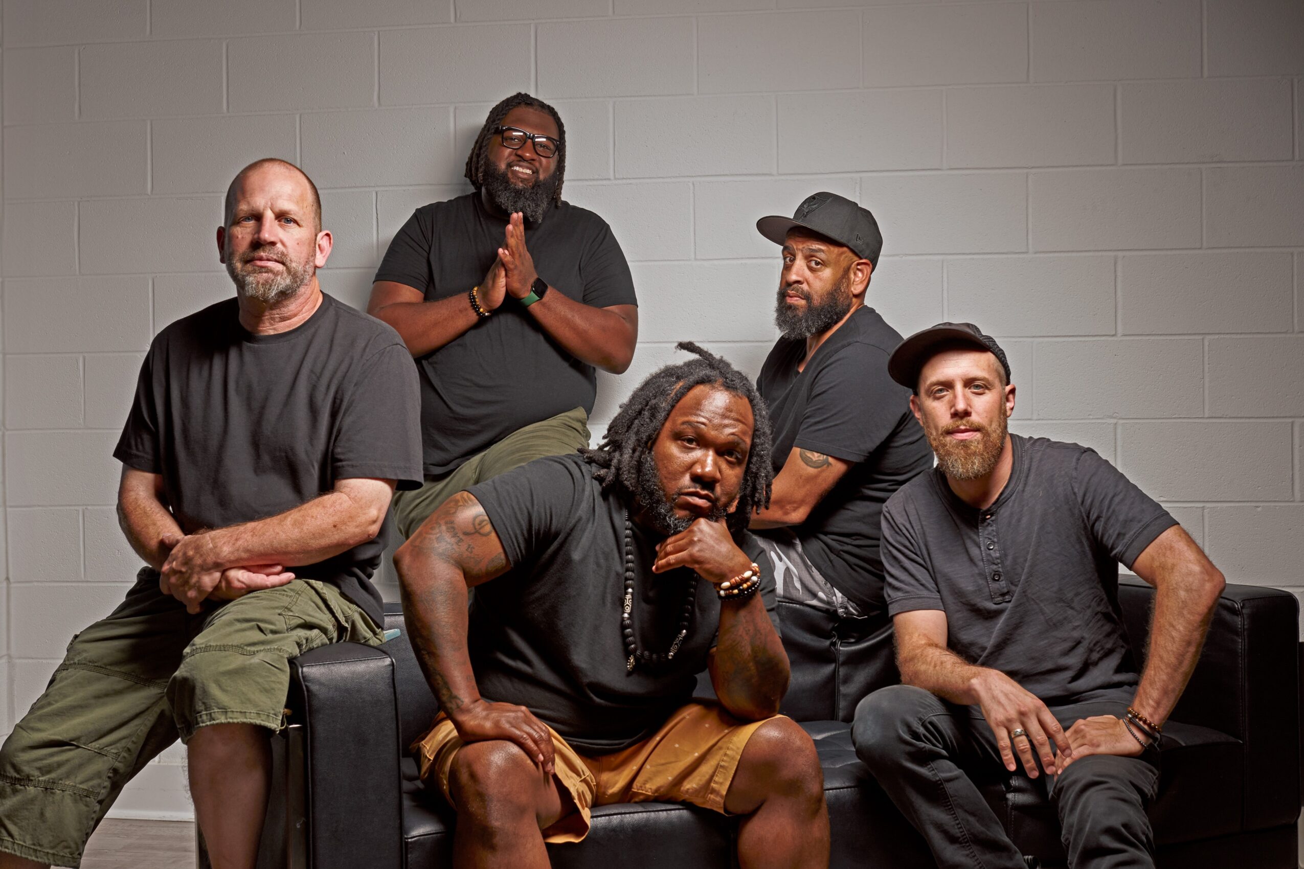 Five men sitting and standing against a gray brick wall, some with hands clasped or resting, wearing casual clothing.