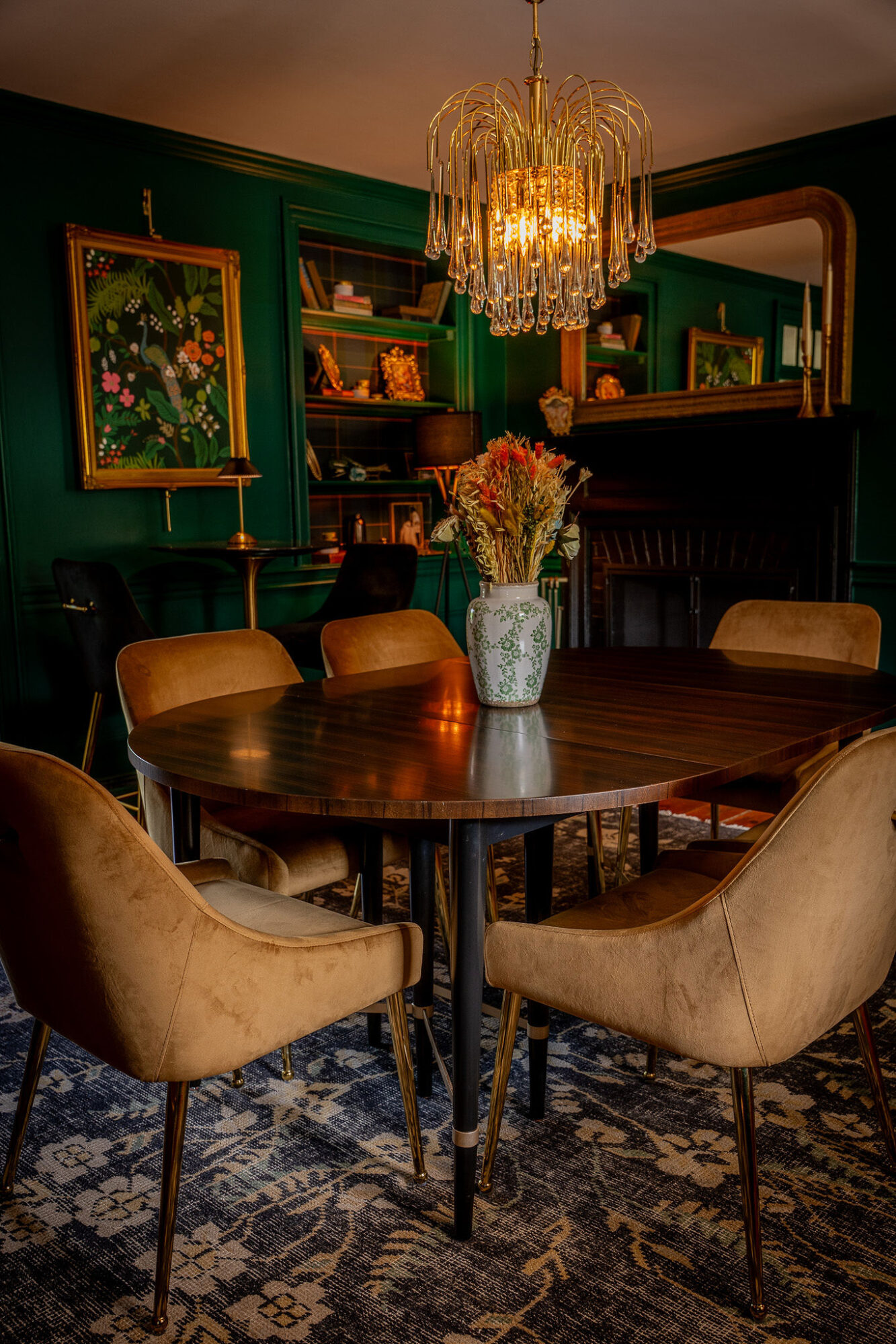 Dining room with a round table, six beige chairs, a chandelier, green walls, and a bookshelf with decorative items.