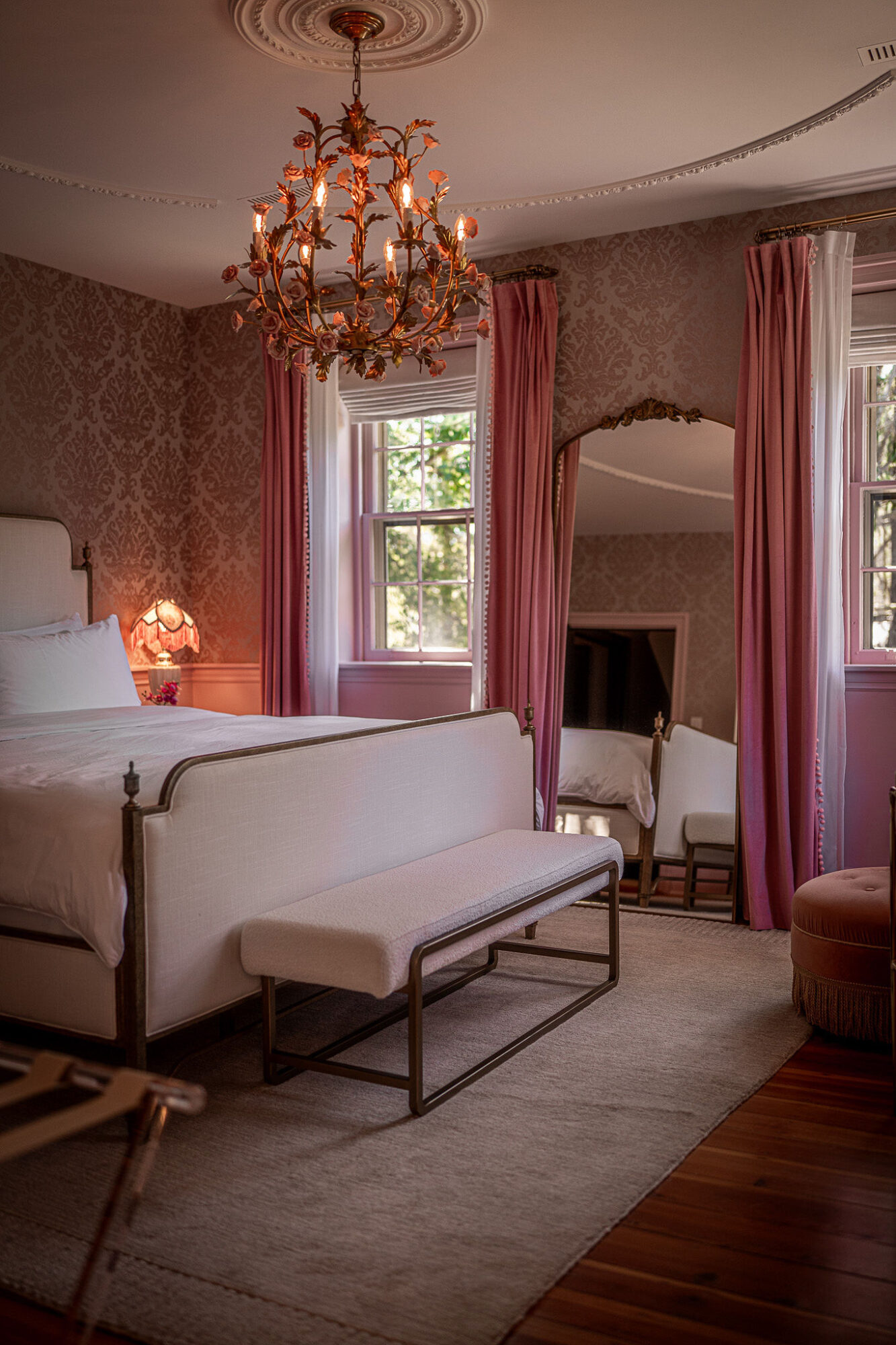 Elegant bedroom with chandelier, pink curtains, white furniture, and a large window with greenery outside.