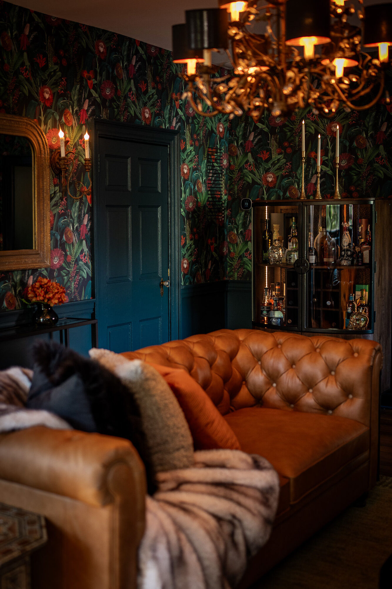 Living room with dark floral wallpaper, blue door, leather sofa, chandelier, and glass cabinet, dimly lit.