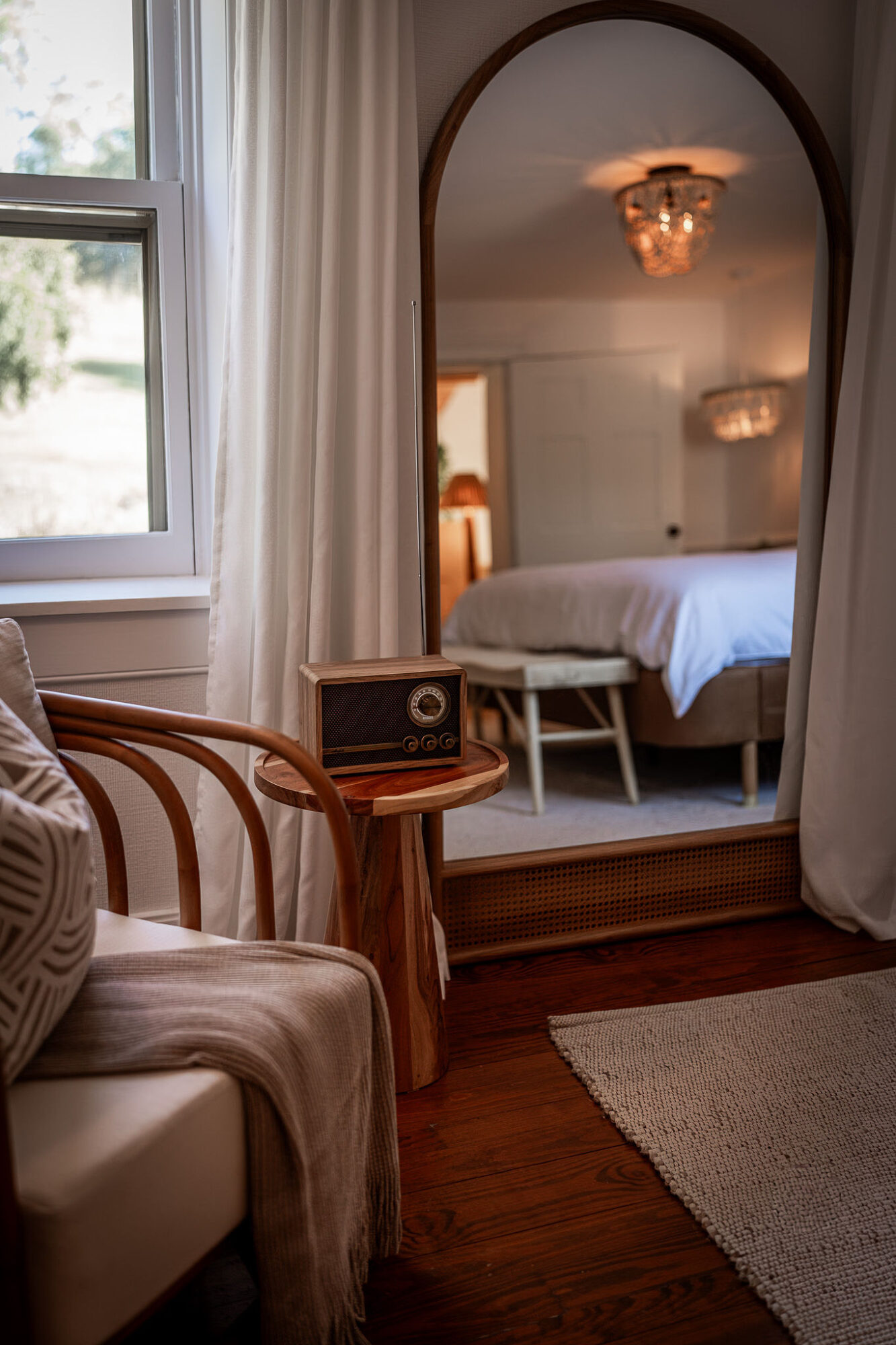 View through an archway into a bedroom with a bed, nightstand, and a chair, seen from a sitting area with a window and curtains.