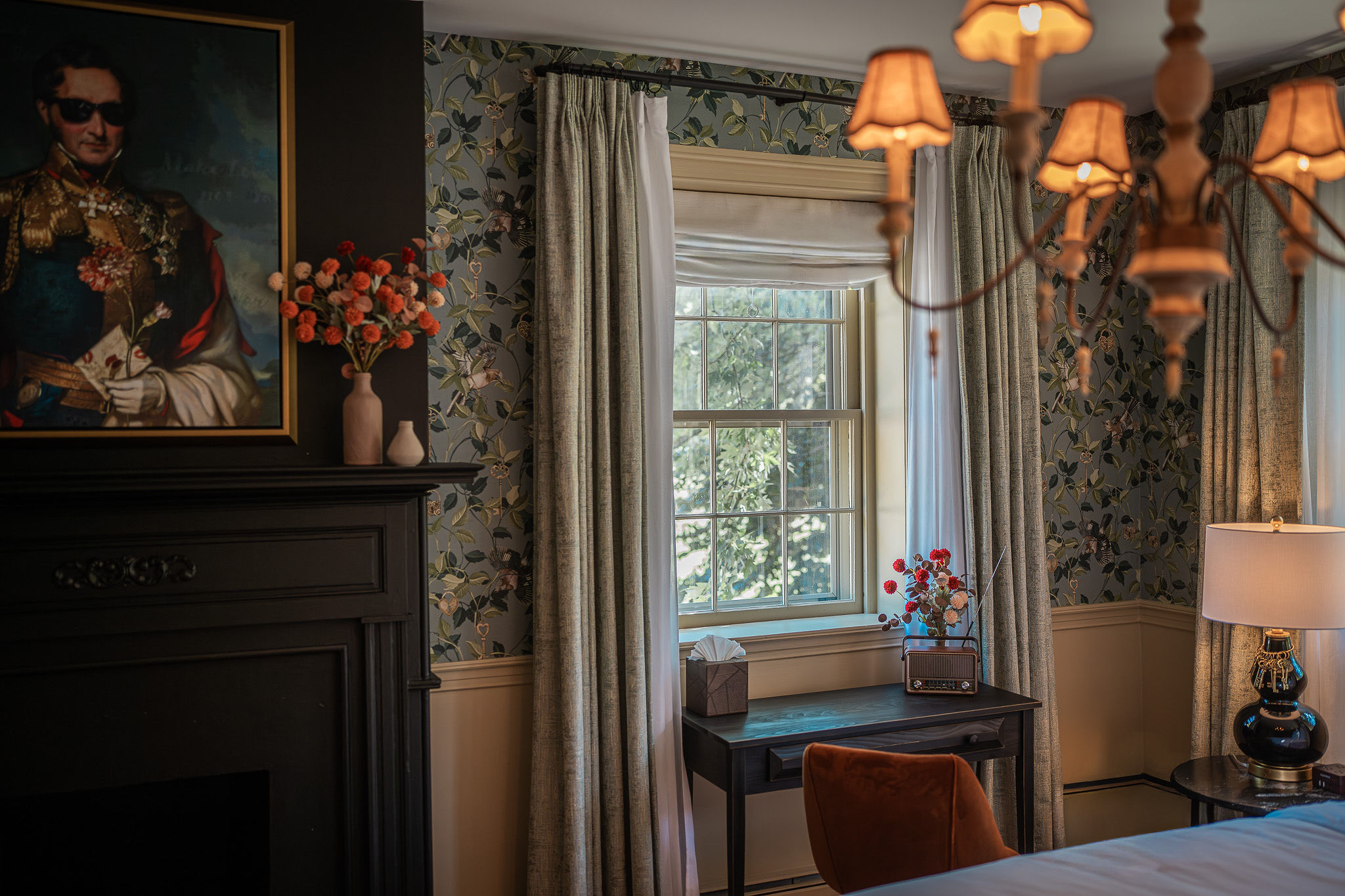 Interior room with window, floral wallpaper, curtains, chandelier, and a table with flowers, a lamp, and a painting on the wall.
