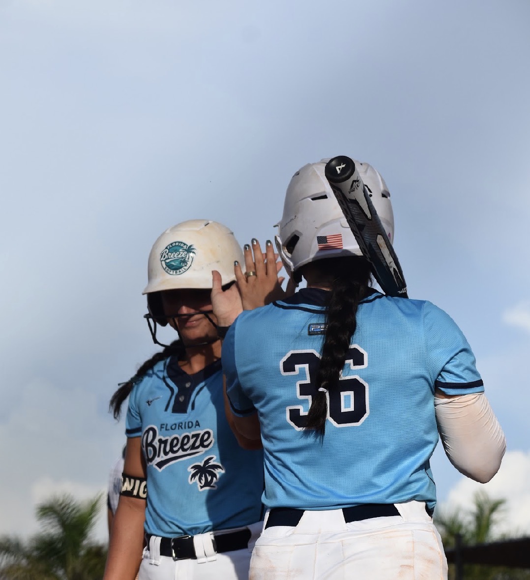 Two female softball players in blue jerseys and white helmets high-five outdoors against a blue sky.