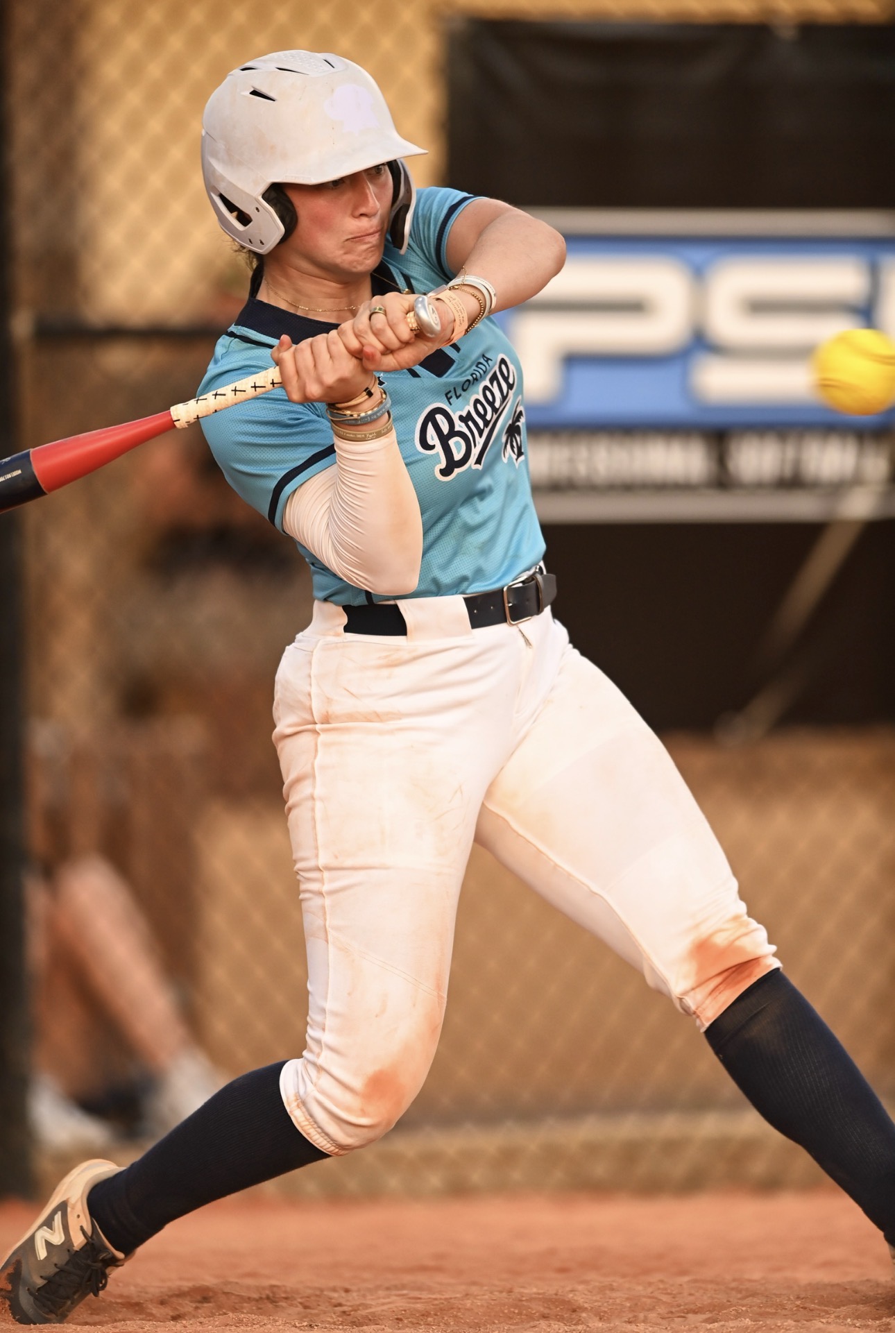 Female softball player swinging a bat, wearing a helmet and uniform, on a dirt field with a fence background.