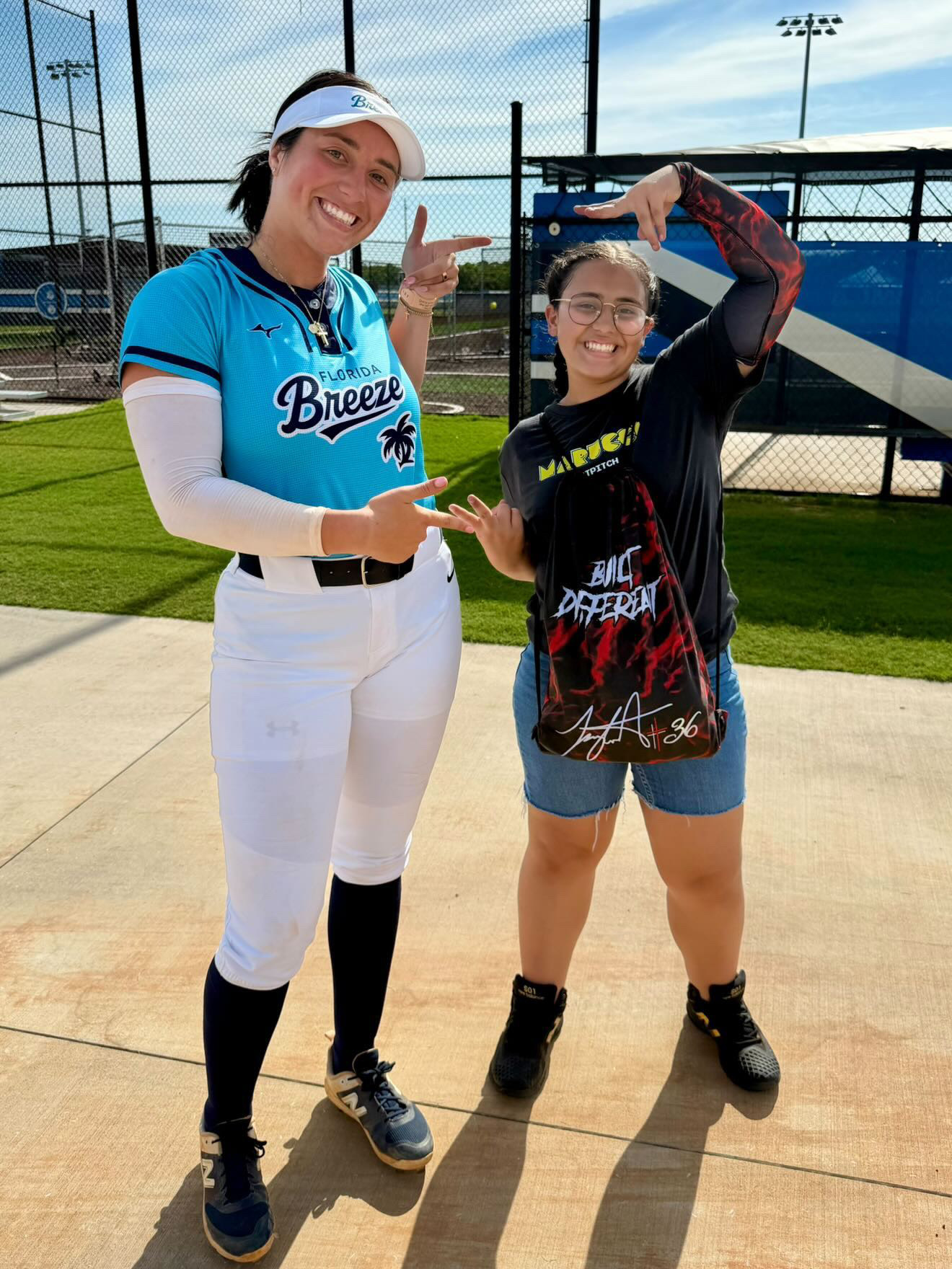 Two women standing on a baseball field, smiling, with a fence and scoreboard in the background.