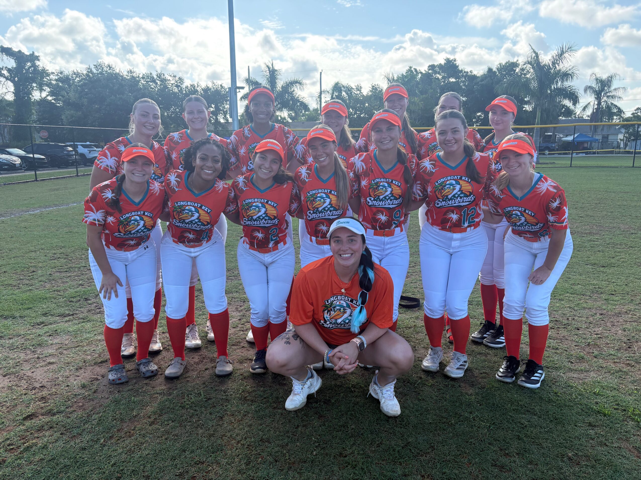 Group of female softball players in red and white uniforms on a field, with trees and a cloudy sky in the background.
