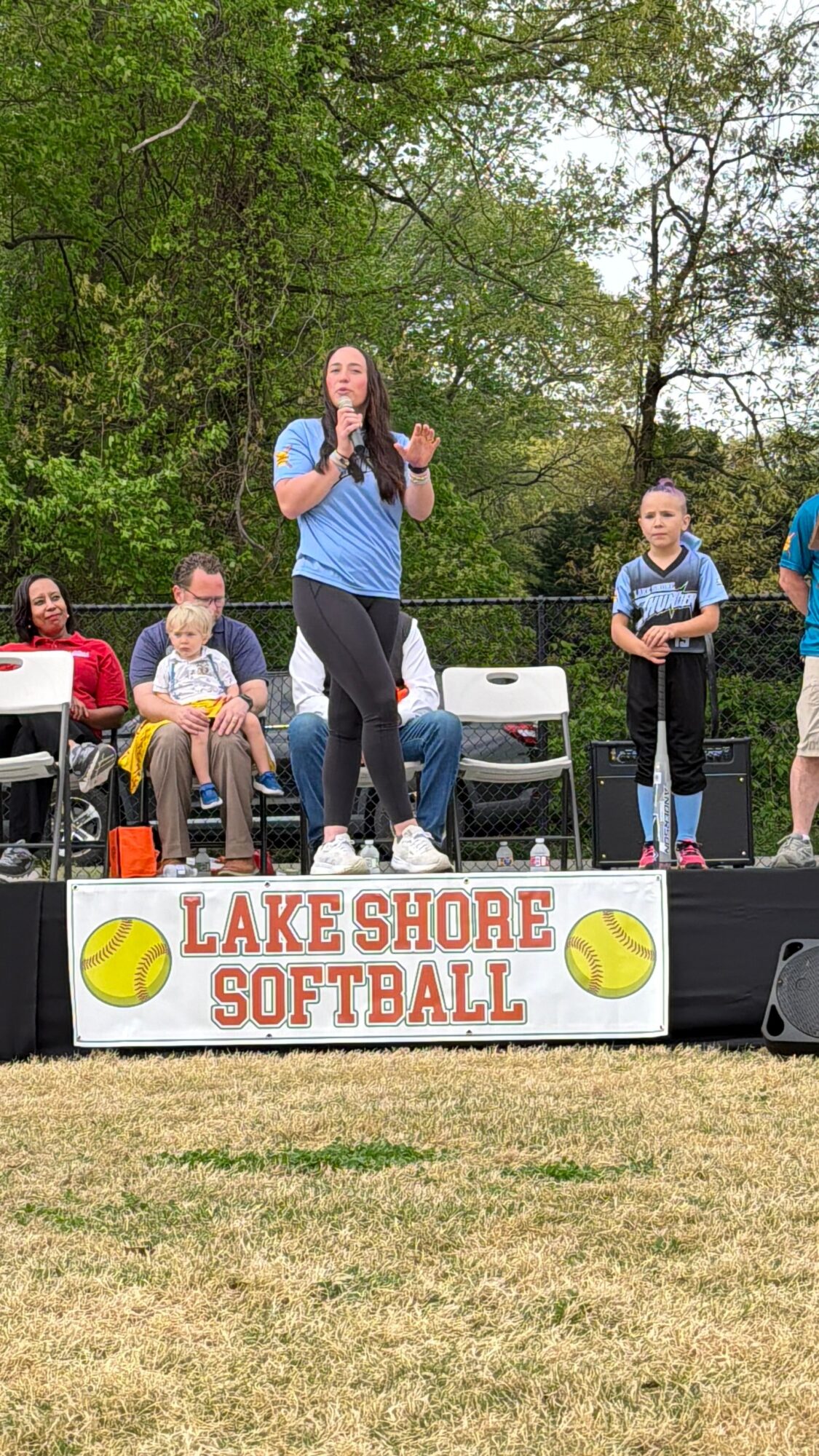 A woman speaking on stage at Lake Shore Softball event with children and adults around, outdoors with trees in background.