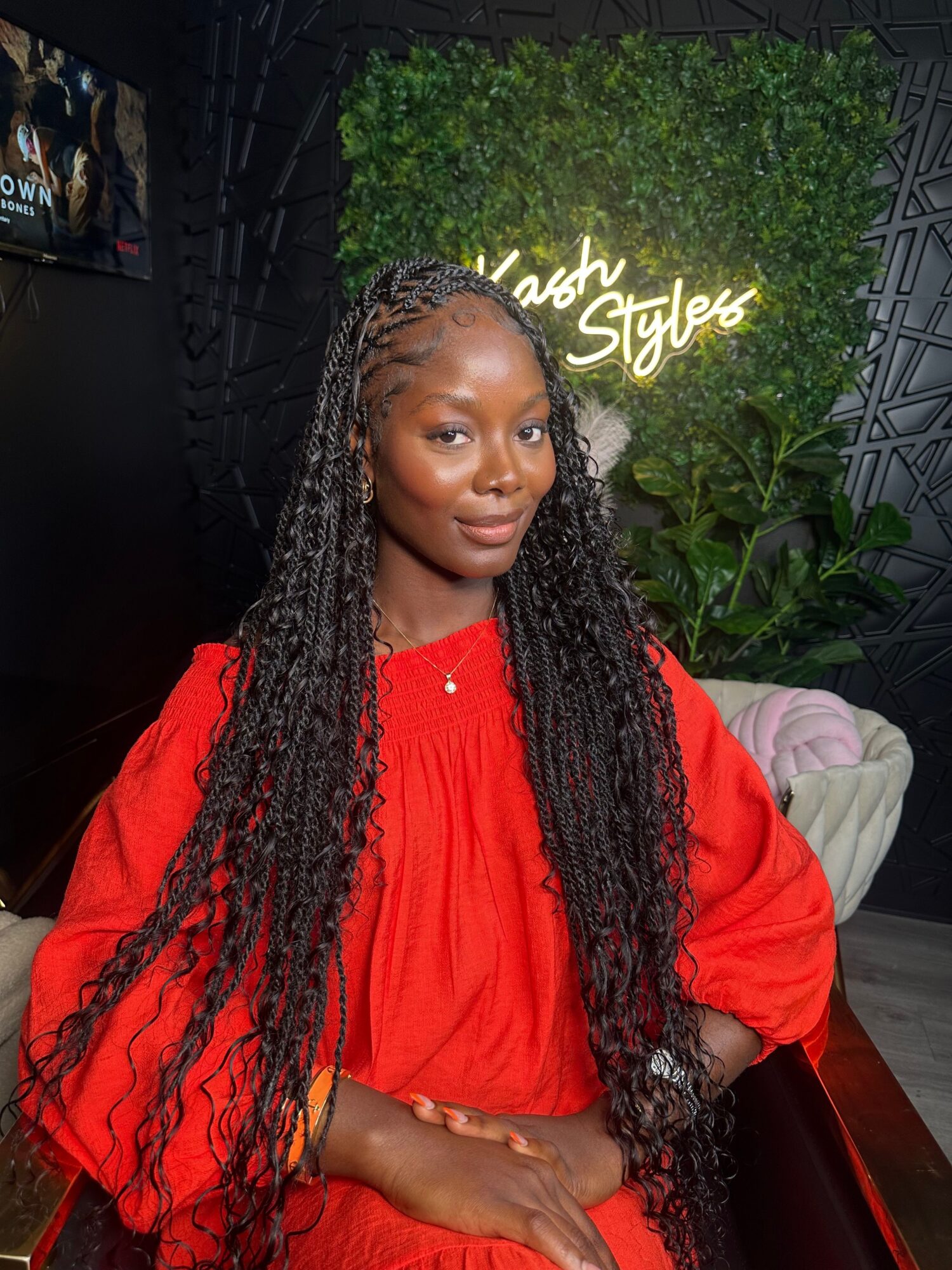 Woman with long braided hair wearing a red top, sitting indoors with a green wall and sign in background.