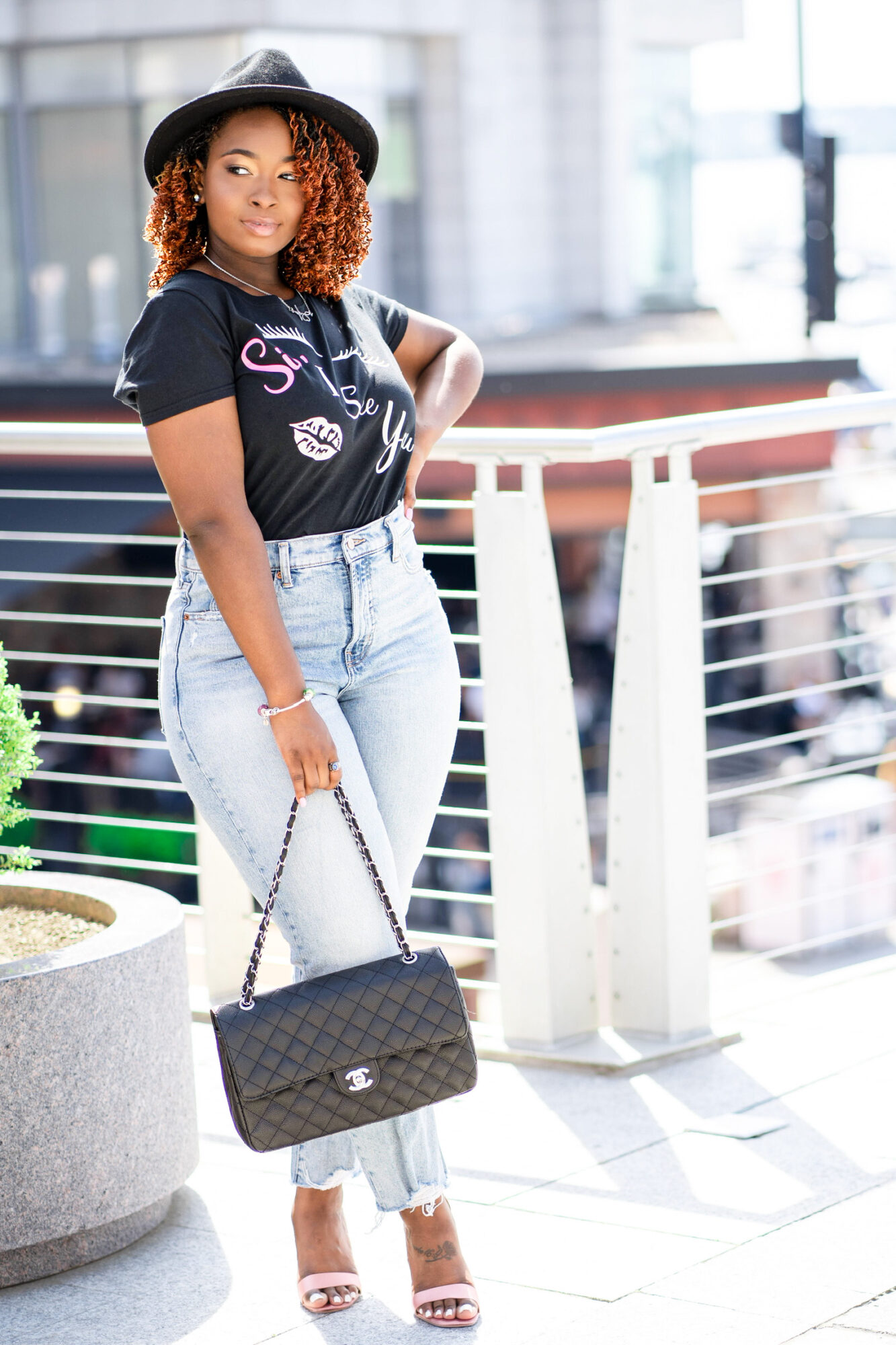 Young woman with curly hair wearing a hat, black t-shirt, light jeans, and heels, holding a black purse outdoors.