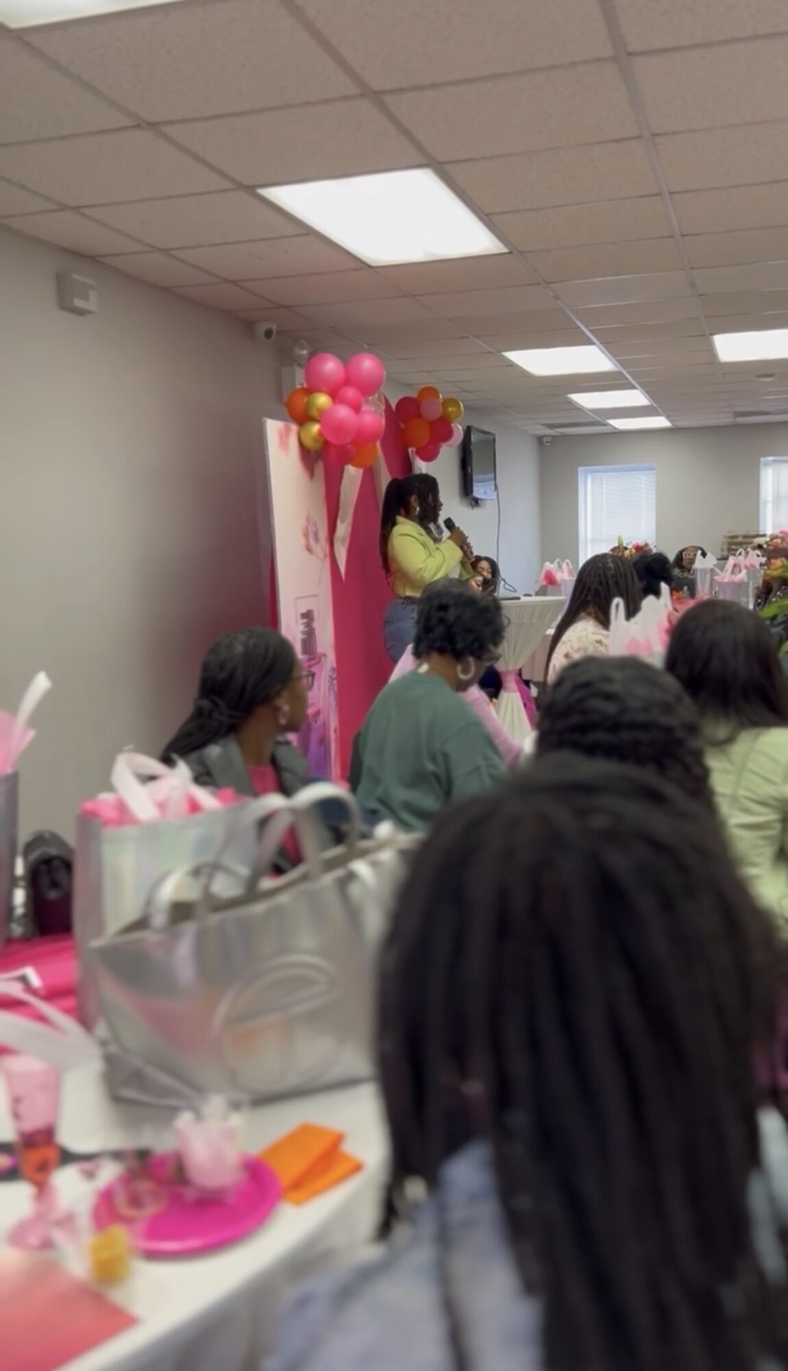 People gathered in a decorated room with pink balloons and a woman speaking at a podium, celebrating an event.