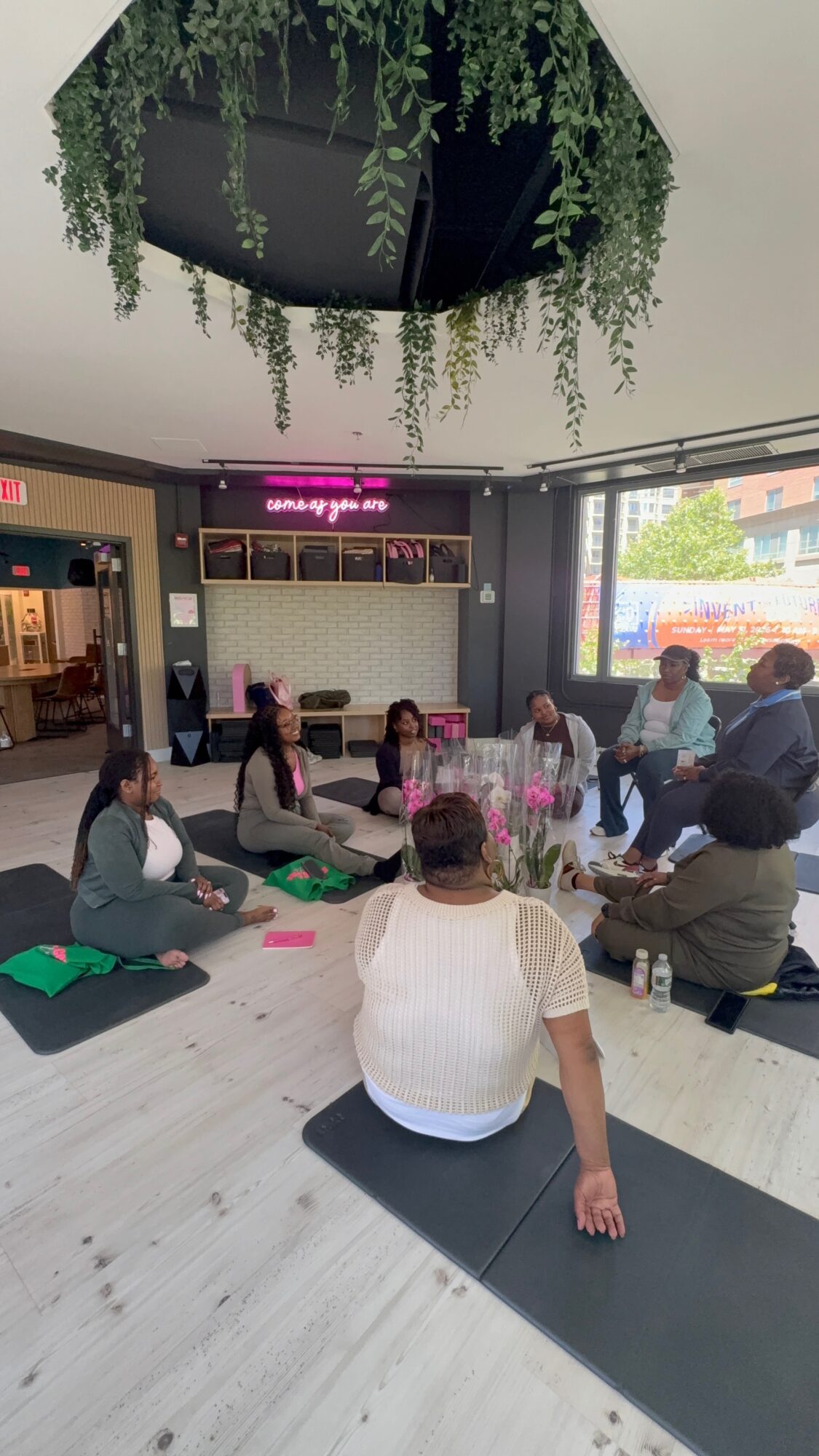 Group of people sitting on mats in a circle inside a room with large windows and hanging greenery from the ceiling.
