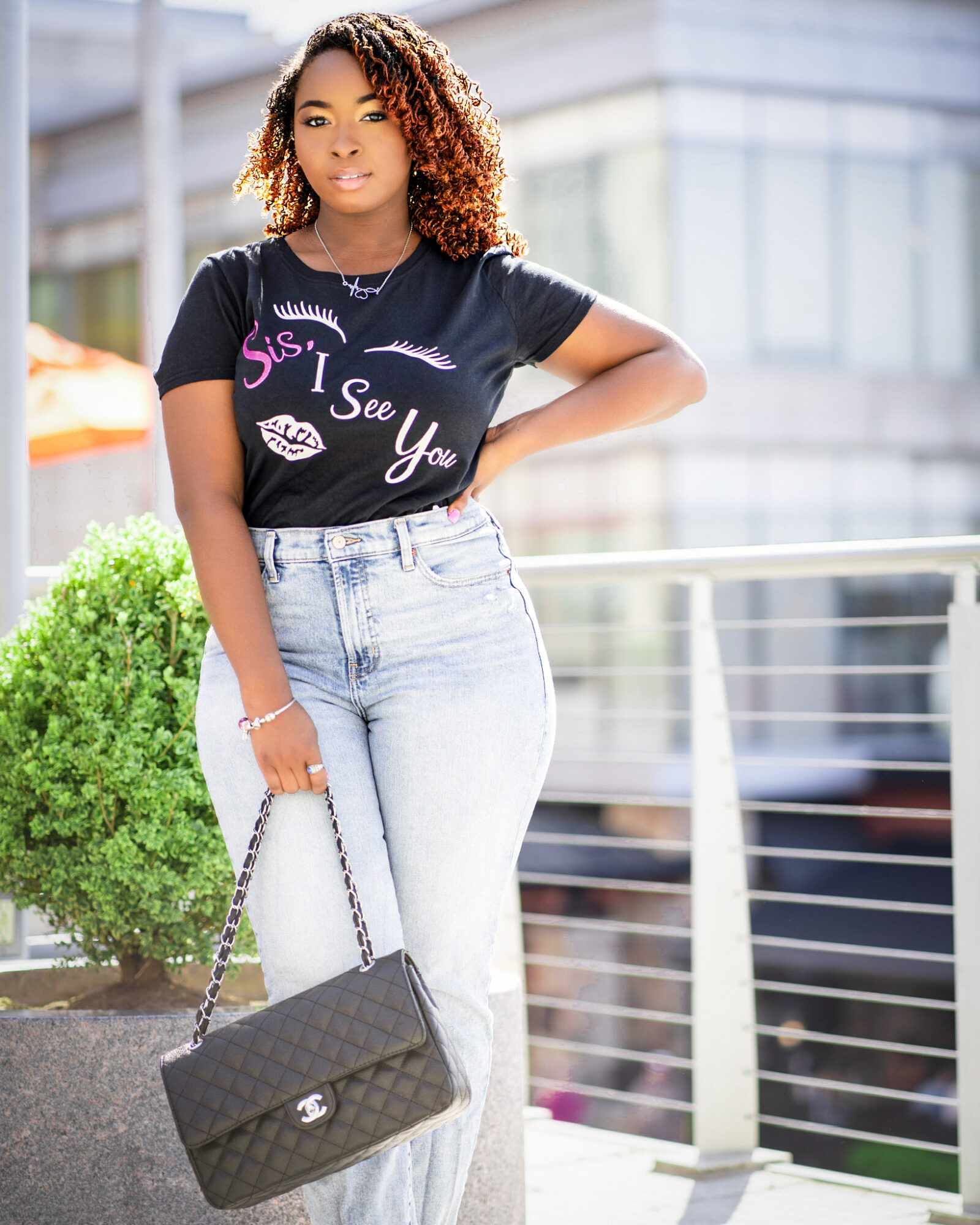 Young woman with curly hair wearing a black T-shirt and light jeans, holding a black purse, standing outdoors near a railing.