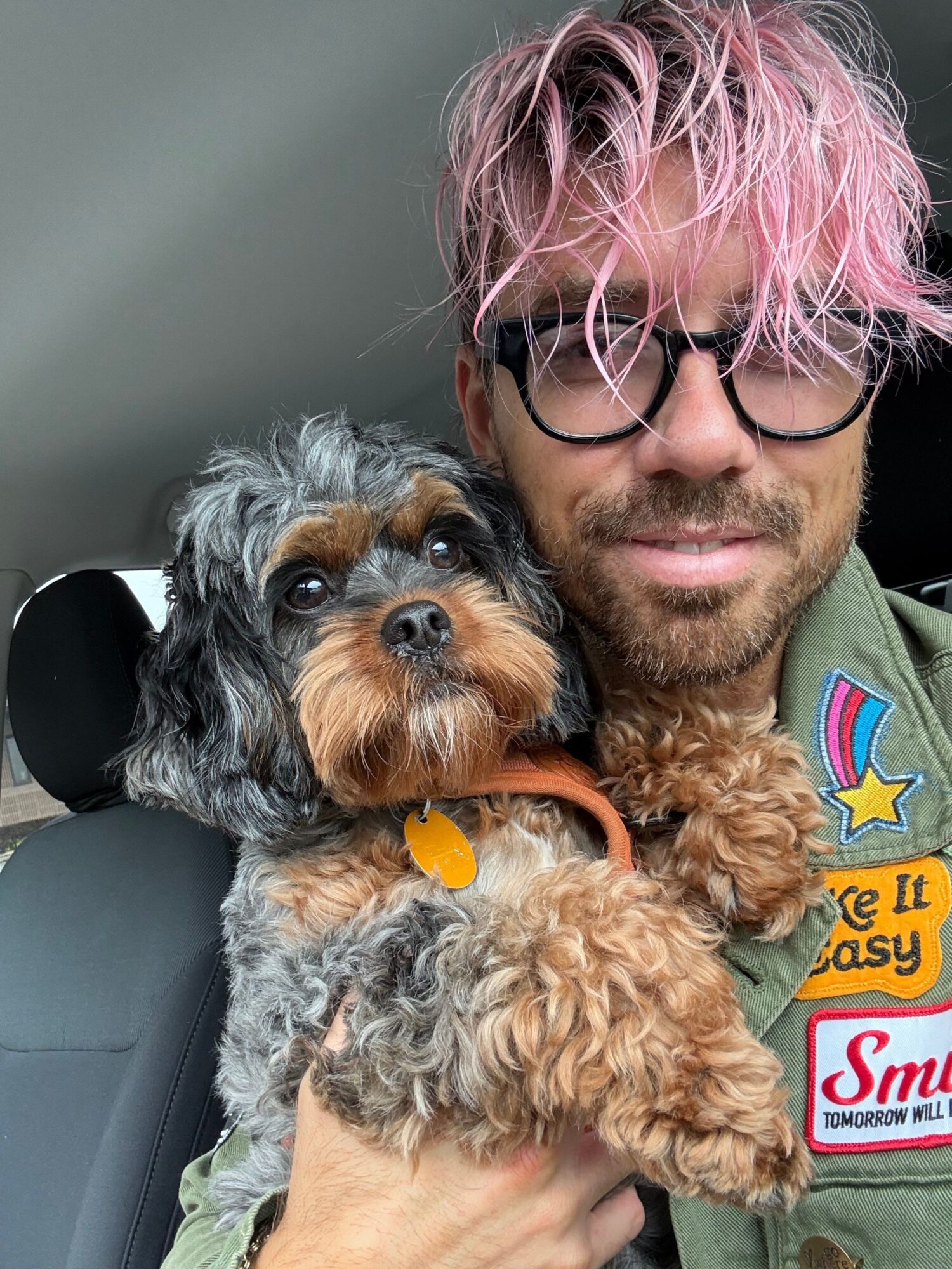 Person with pink hair and glasses holding a small, curly-haired dog inside a vehicle.