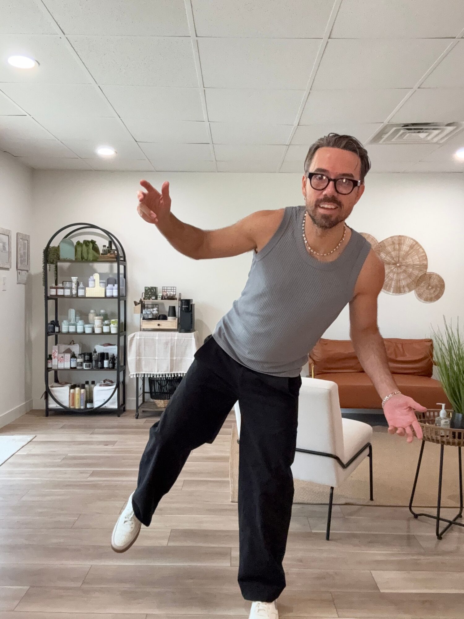 Person standing in a room with white walls, wooden floor, and furniture, smiling and gesturing with hands.