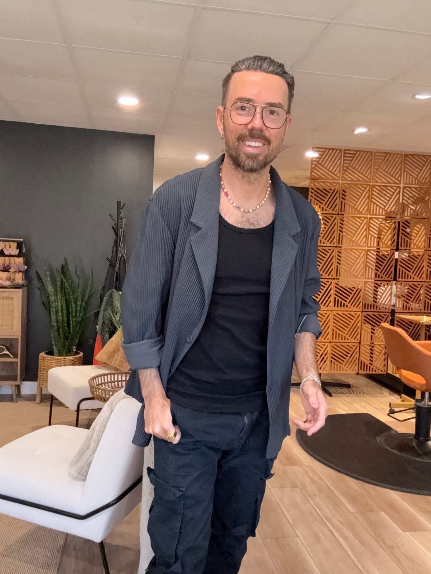 Man with glasses, beard, and short hair standing indoors, wearing a dark shirt and jacket, with a wooden wall and plants behind him.