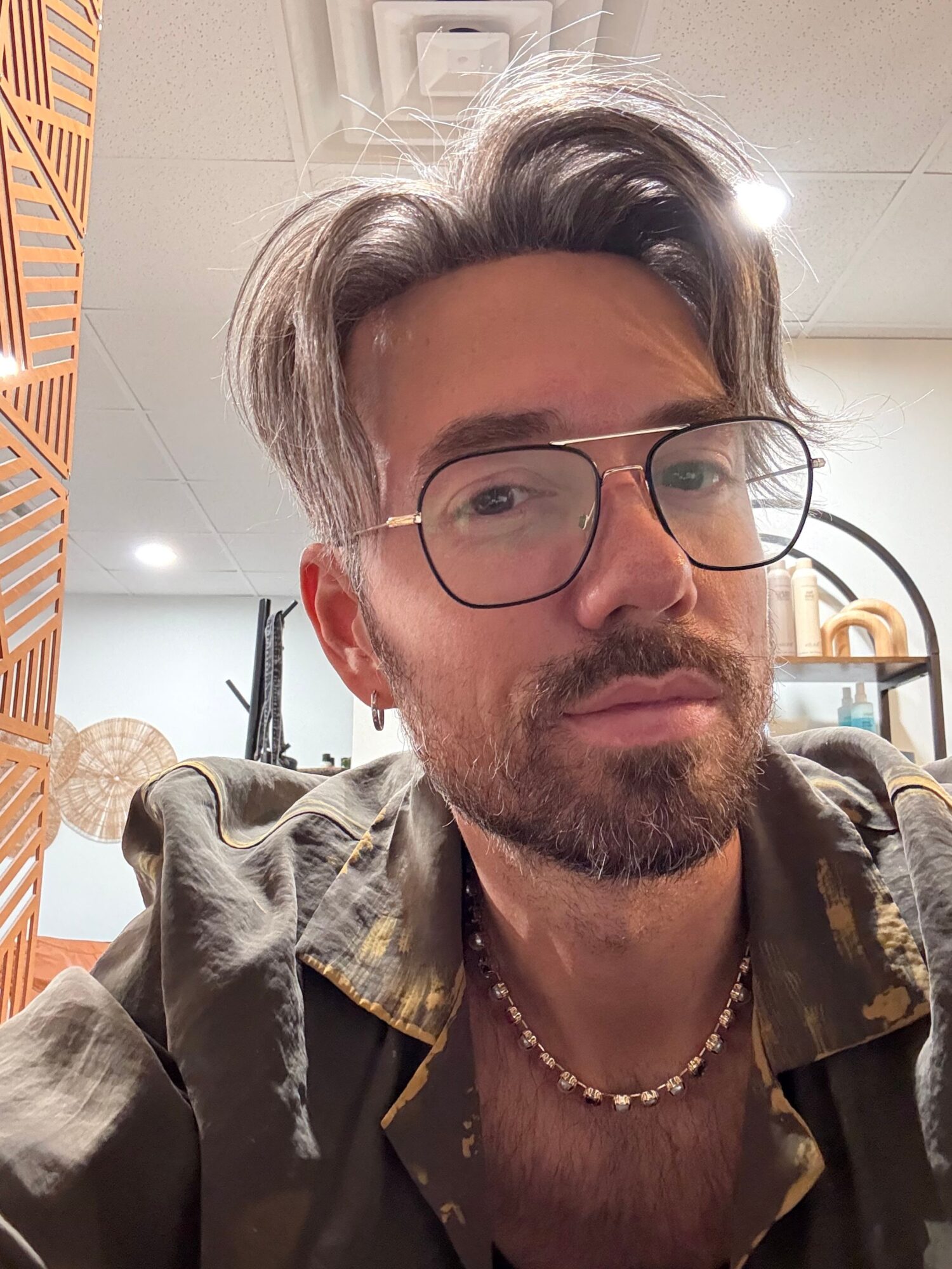 Man with glasses, beard, and long hair, wearing a patterned shirt and necklace, sitting indoors with a neutral expression.