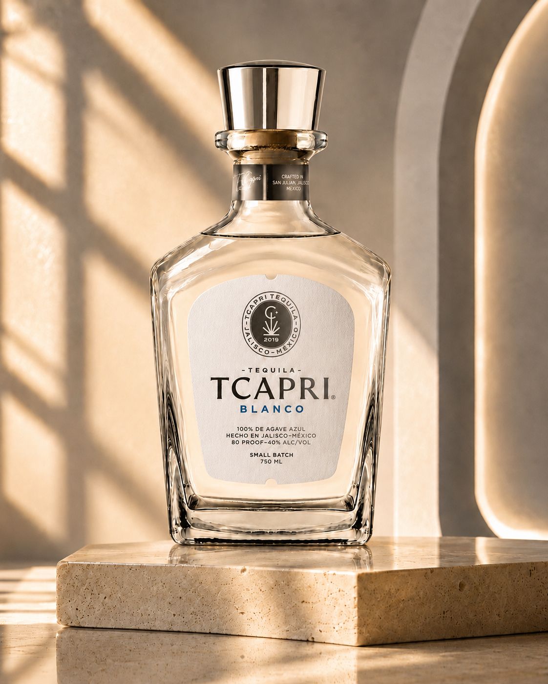 A clear glass bottle of TACPRA Blanco perfume with a silver cap, placed on a beige surface with sunlight and shadows.