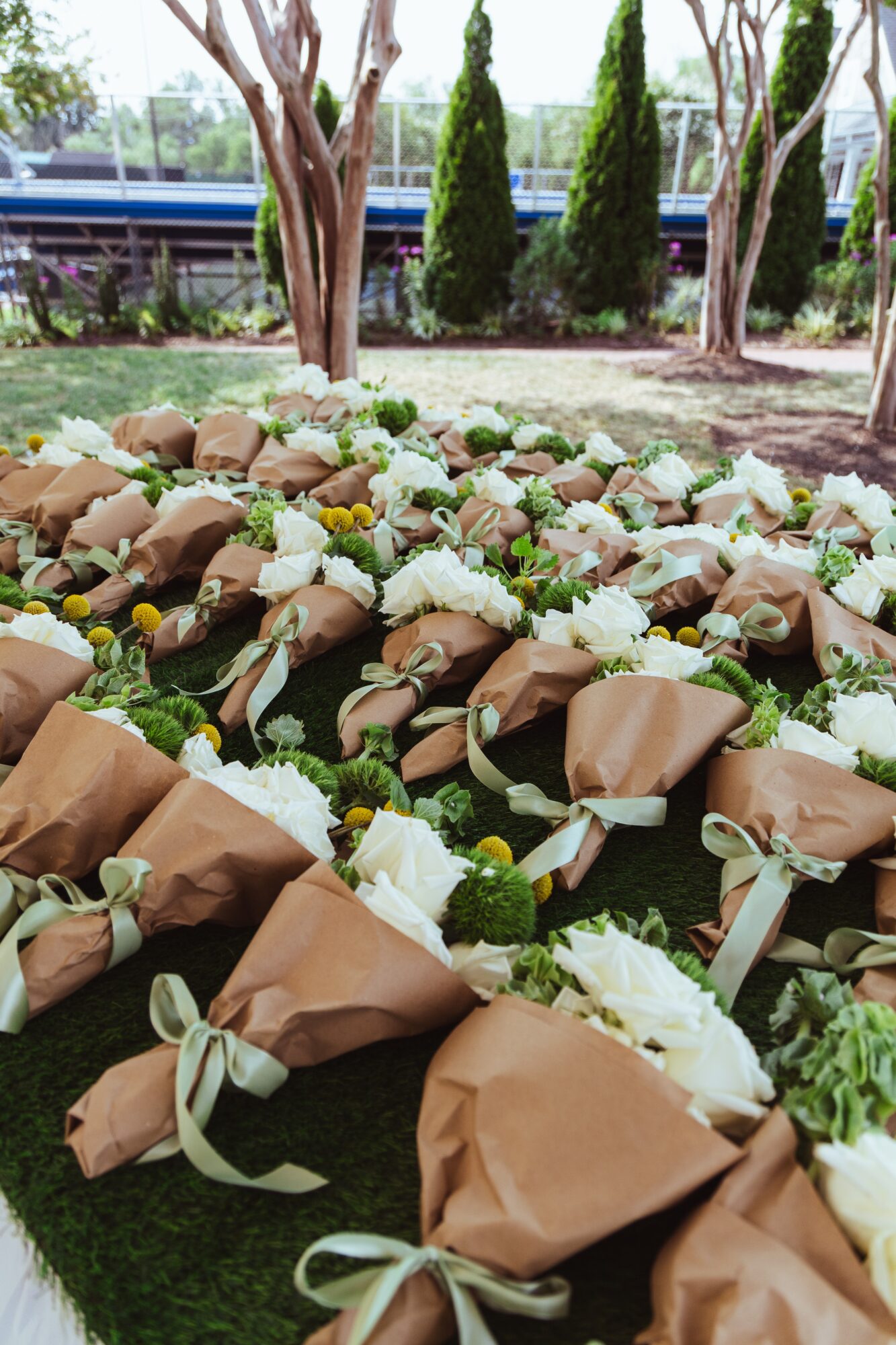Multiple flower bouquets wrapped in brown paper and tied with light-colored ribbons arranged on grass outdoors.