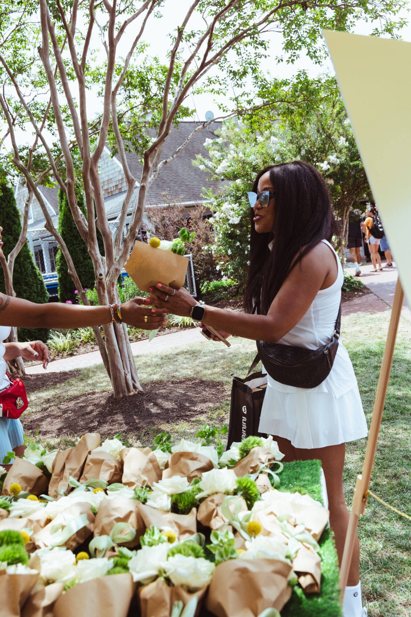 Woman in white dress and sunglasses receives a coconut from another person outdoors, with trees and people in background.