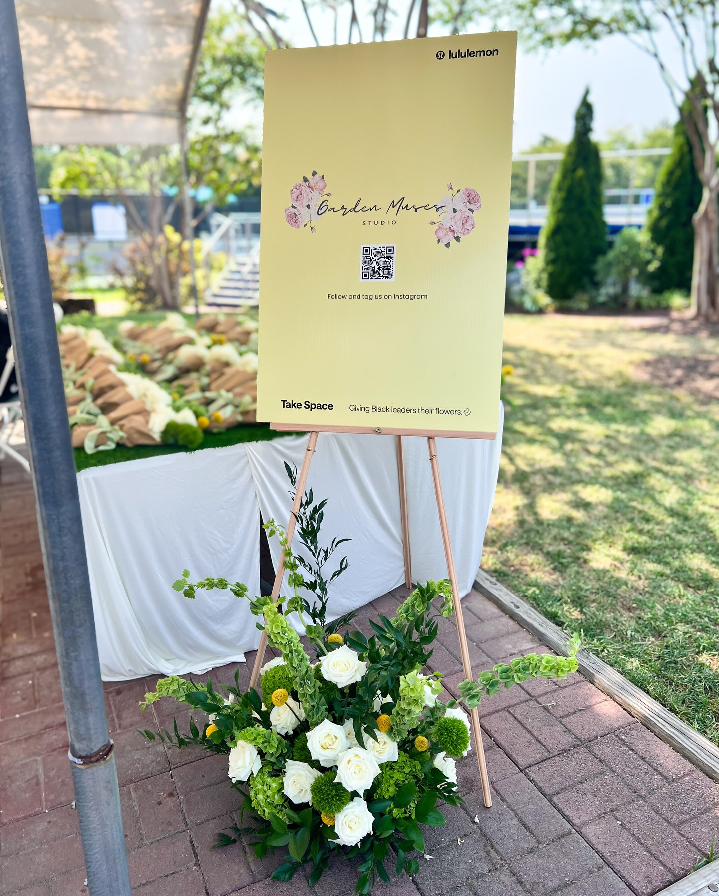 Signboard with floral design and QR code, placed on a stand in a garden setting, with a flower arrangement at the base.