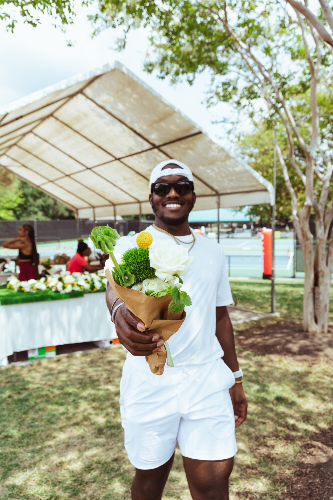 Smiling man holding a bouquet of white and green flowers outdoors, with a table and trees in background.