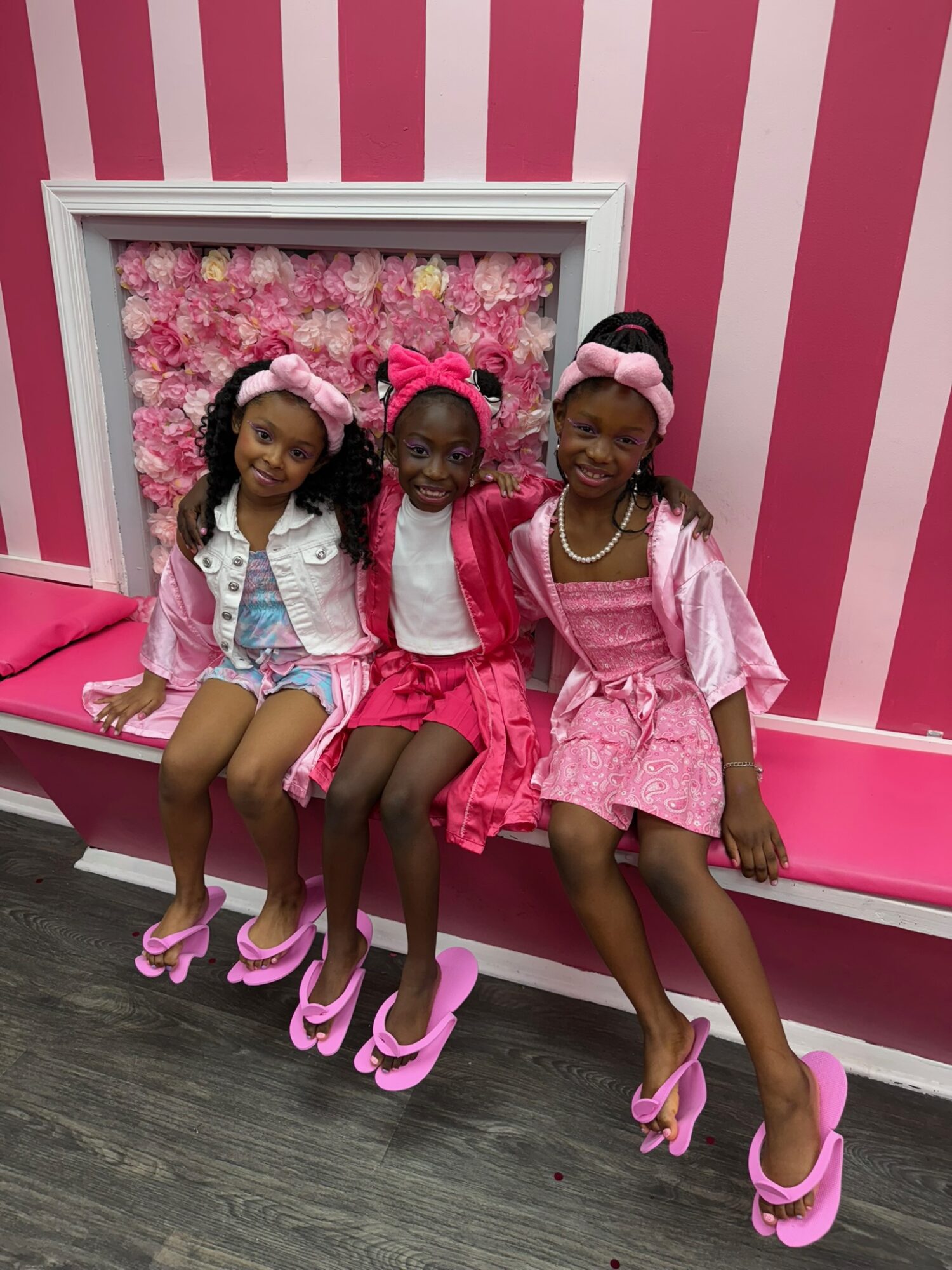 Three young girls sitting on a pink bench with a floral backdrop, wearing pink dresses and matching pink shoes, smiling.