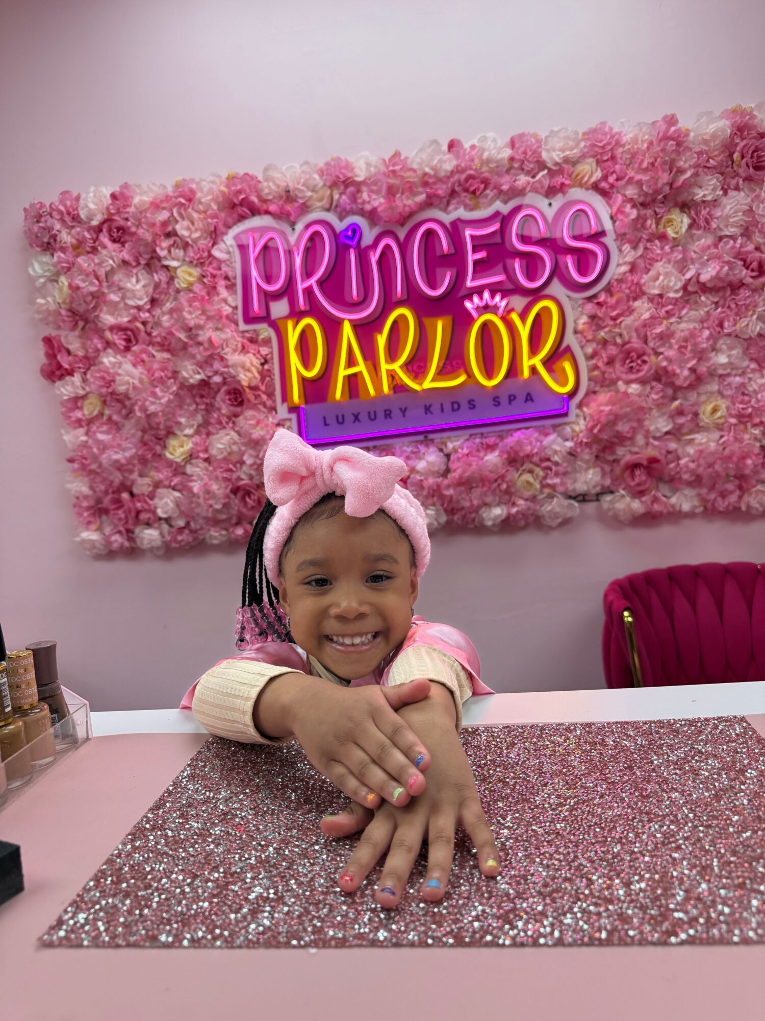 Child with pink headband and bow, sitting at a pink table with pink glitter tablecloth, in front of a pink floral backdrop with Princess Parlor sign.