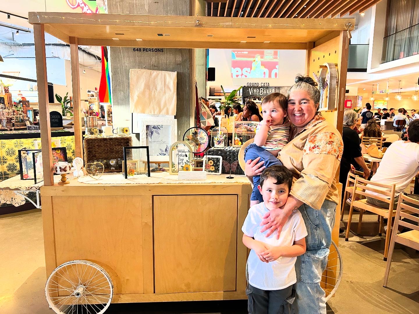 Woman holding a child and a boy standing in front, in a busy indoor market or food court area.