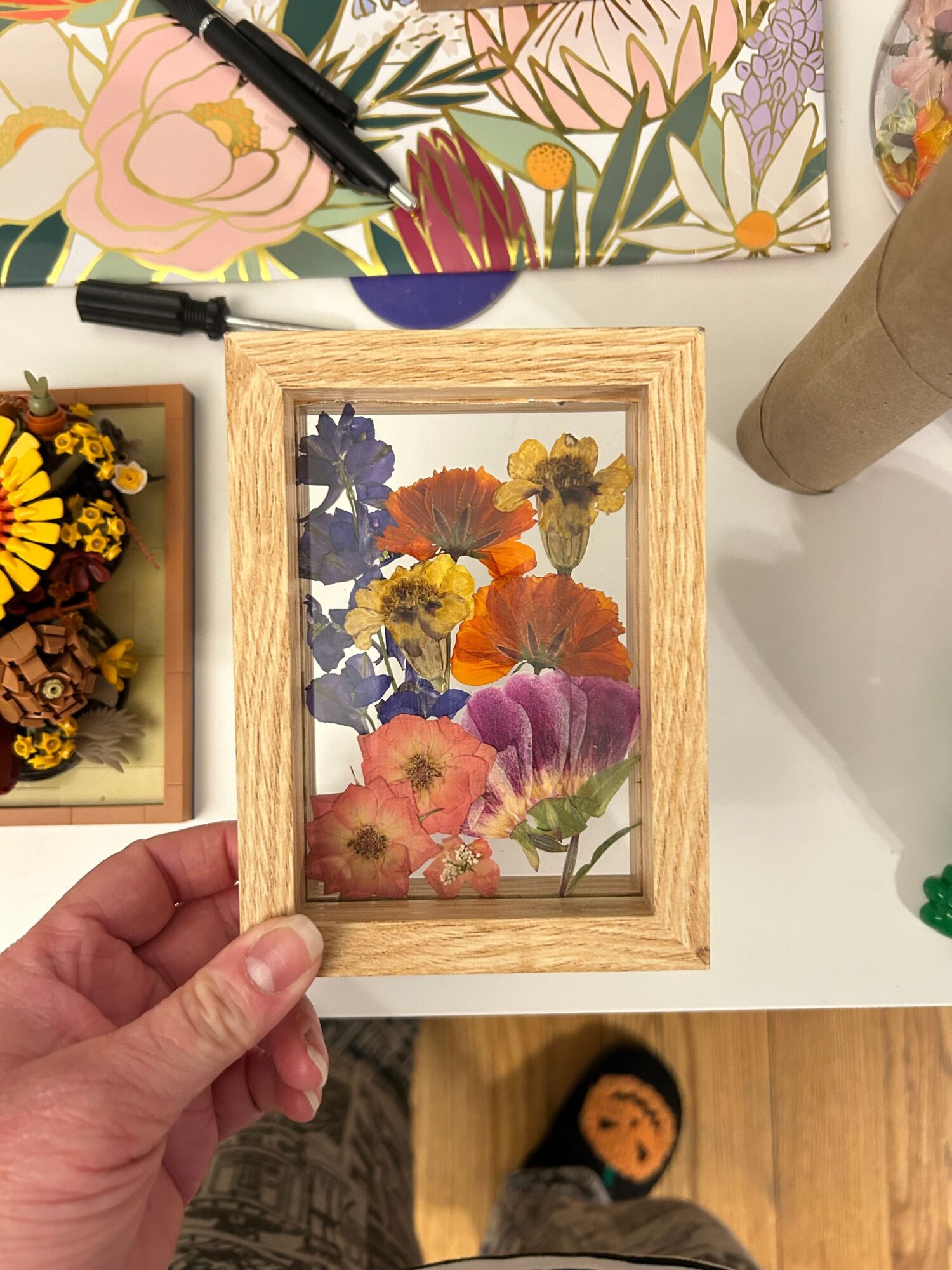 Person holding a framed pressed flower arrangement above a table with more pressed flowers, a marker, and a floral-patterned cloth.