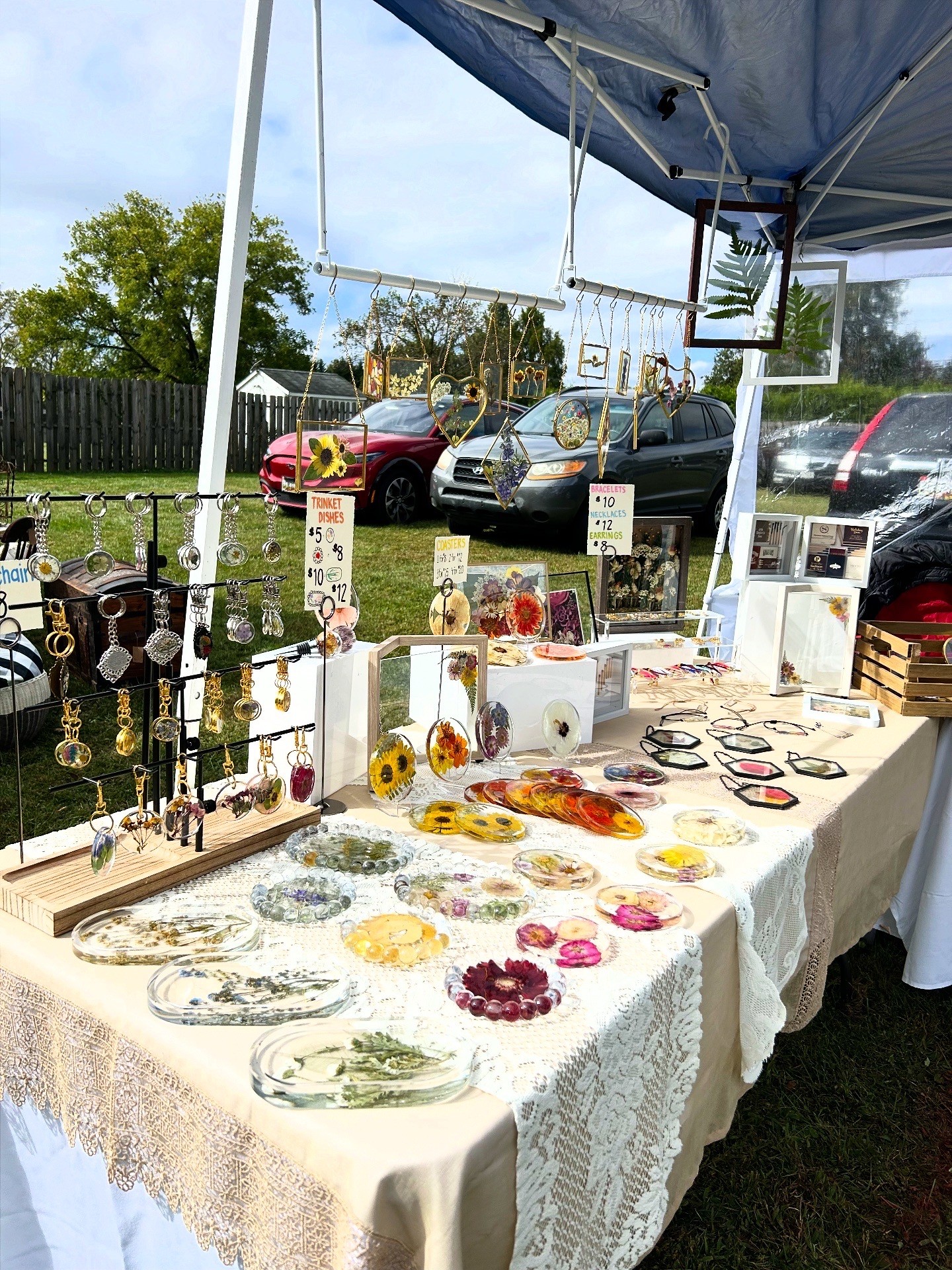 Outdoor market stall displays jewelry and accessories on a table with cars and trees in the background.