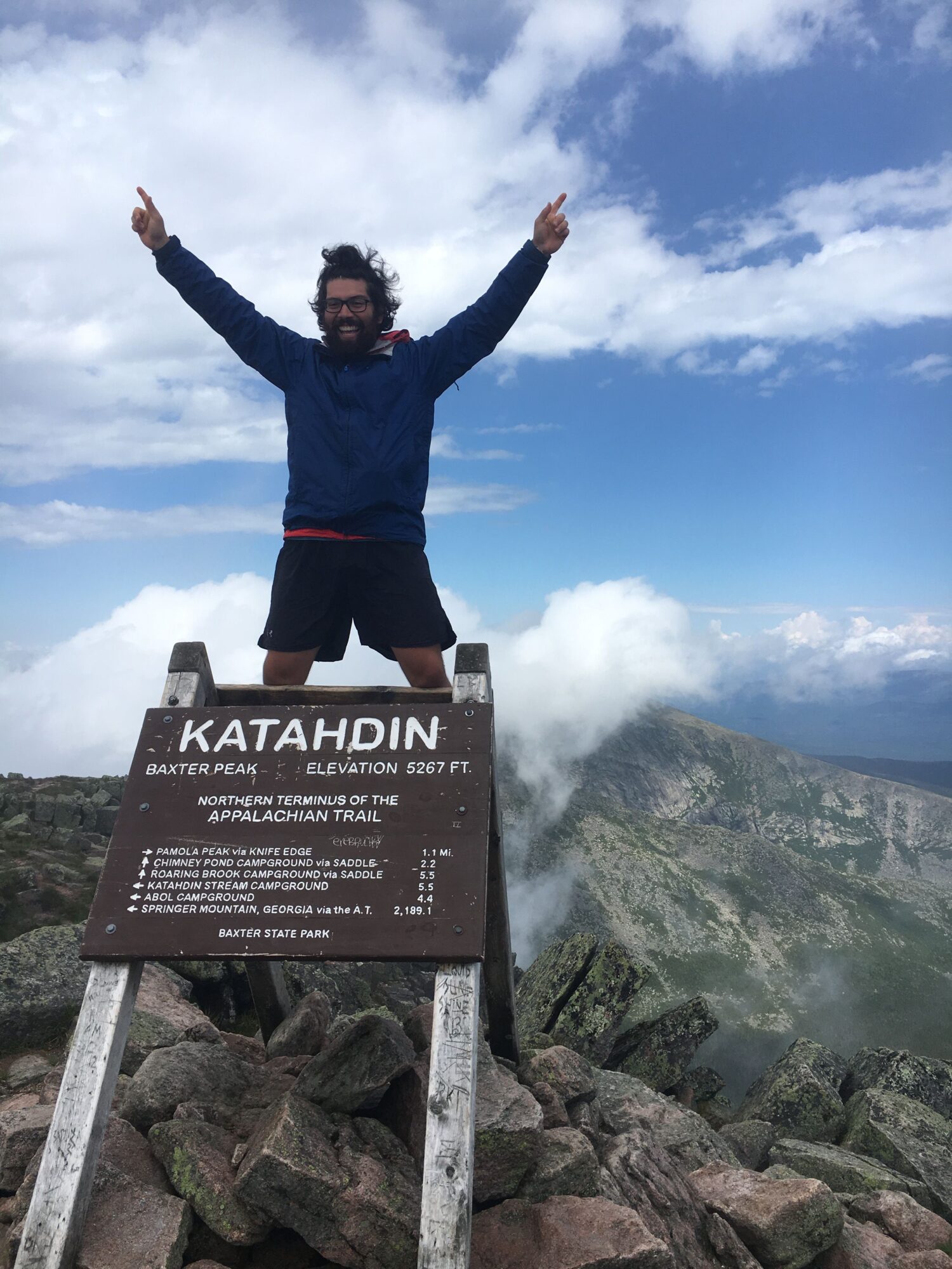 Person standing on rocky mountain peak with arms raised, cloudy sky background, and a signpost with text in a foreign language.