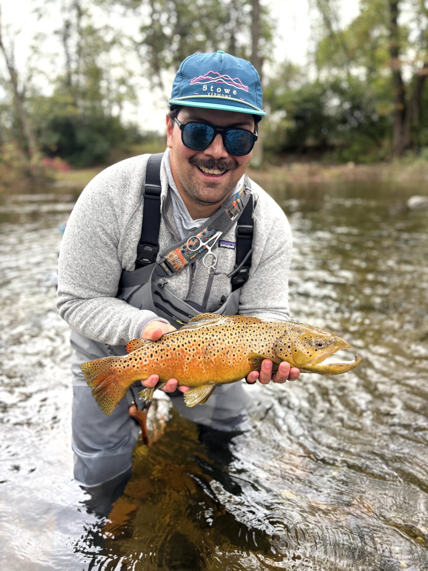 Man in outdoor gear holding a fish in a river, smiling, wearing sunglasses and a cap.