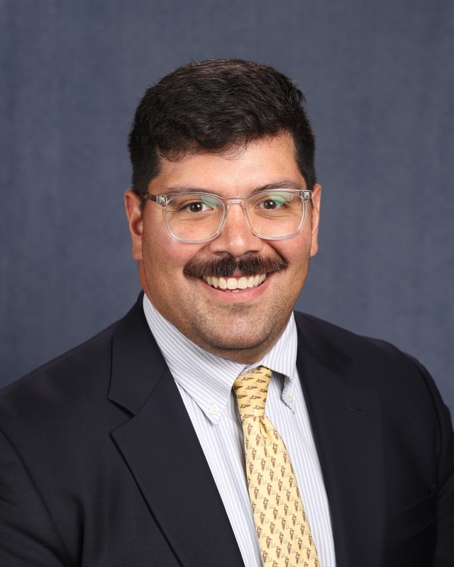 Man with glasses, dark hair, mustache, smiling, wearing a suit, white shirt, and yellow tie, against a plain background.