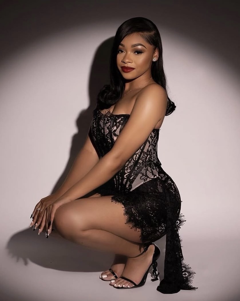 Woman in black lace dress crouching, with dark hair and red lipstick, against a plain background.