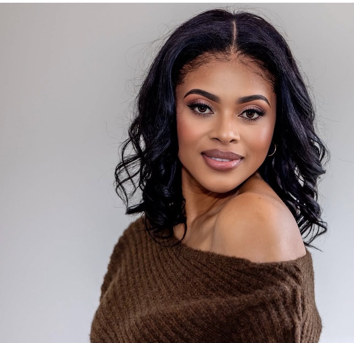 A woman with shoulder-length wavy black hair, wearing a brown off-shoulder sweater, smiling softly against a plain background.