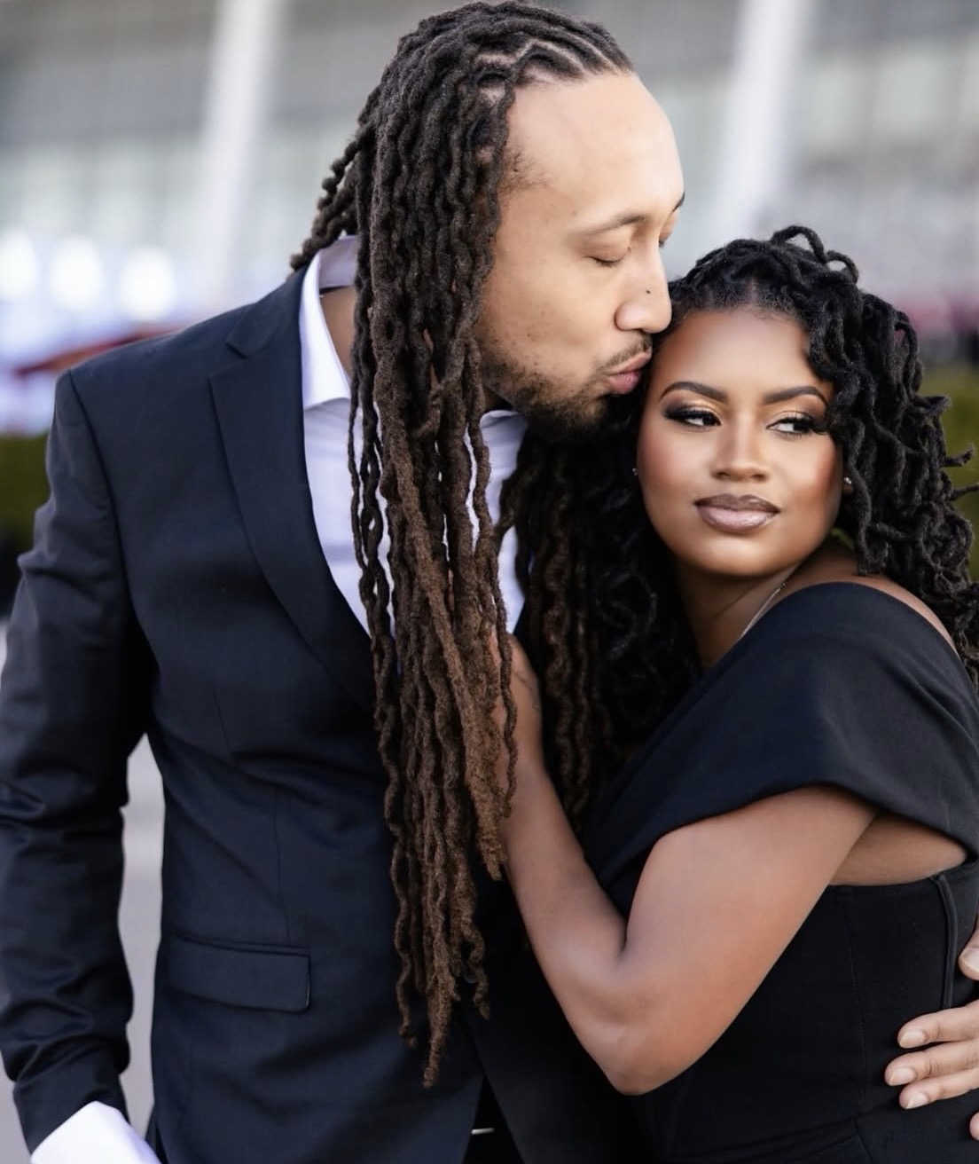 Man with long dreadlocks and woman with curly hair embrace outdoors, close-up shot, both looking serene.